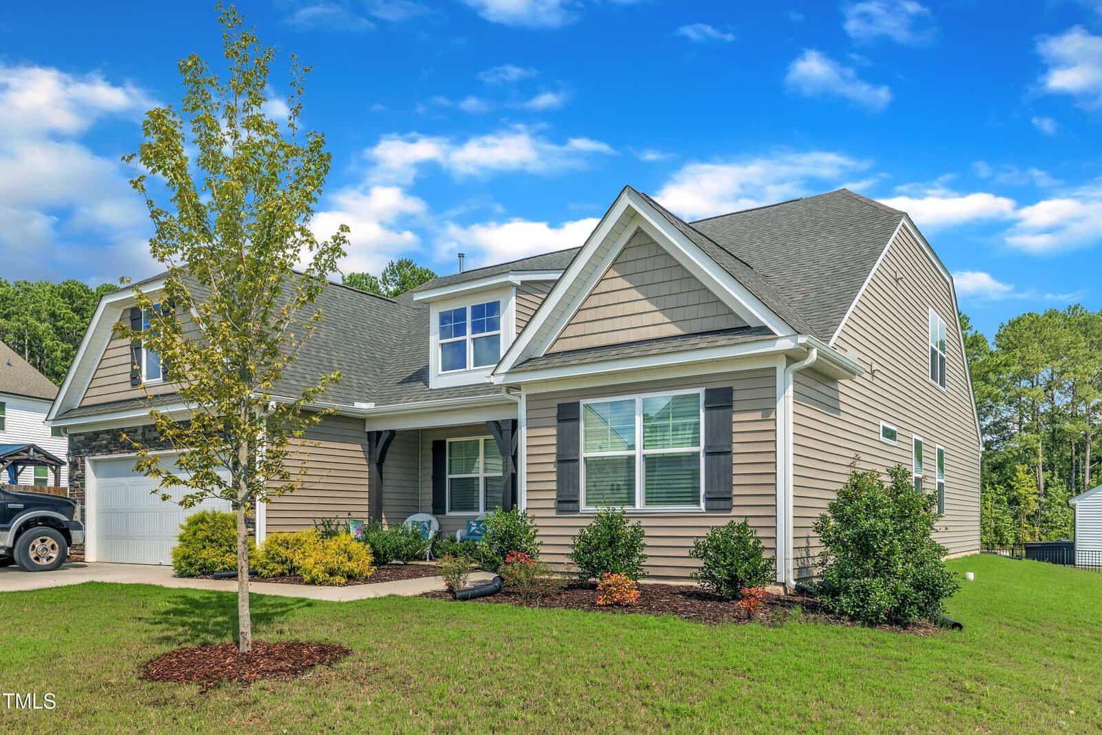A tan two-story suburban house with white trim, black shutters, a front porch, manicured lawn, young tree, and landscaped bushes under a blue sky with scattered clouds, located at 25 Bourne Drive in Franklinton. A black vehicle is partially visible on the left.