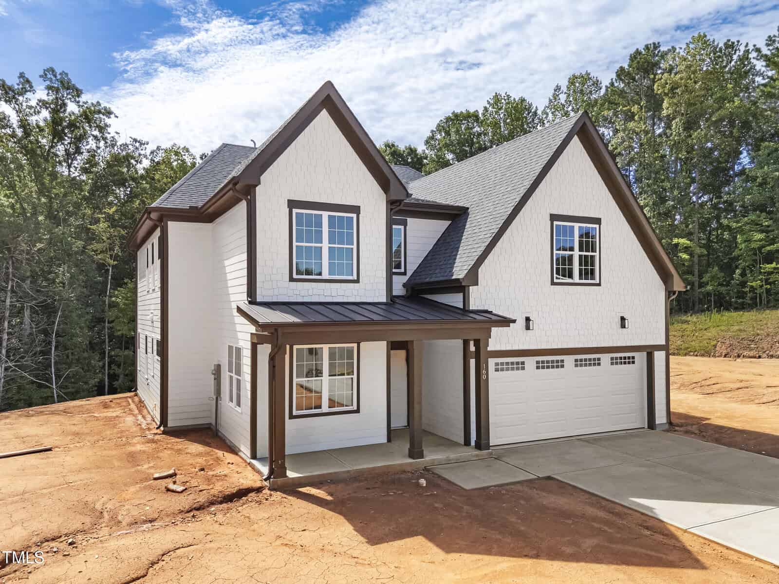 Two-story modern house with white exterior and dark trim, featuring large windows, a covered front entry, and a three-car garage. The house sits on a dirt lot surrounded by trees under a partly cloudy sky.