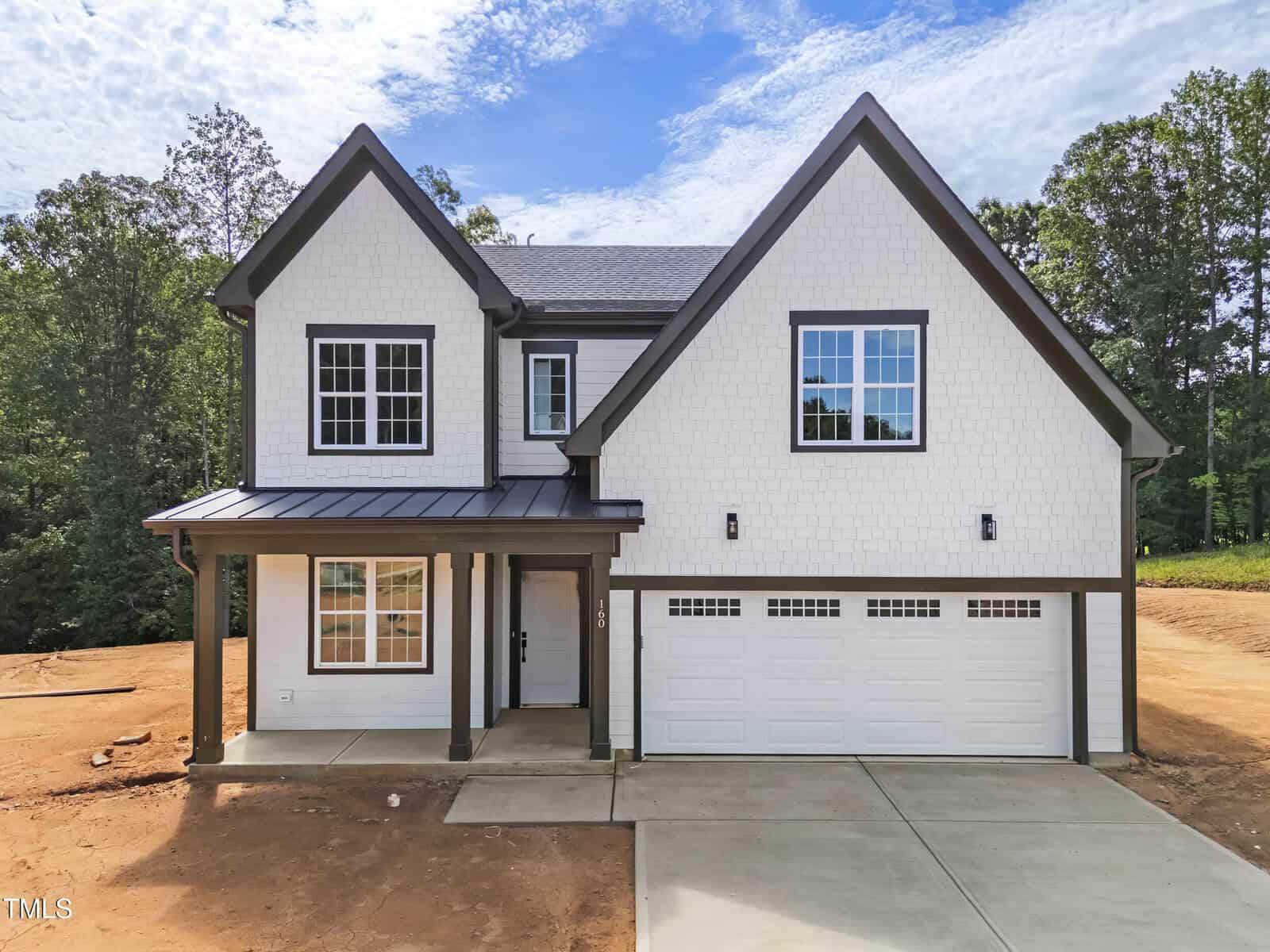 Two-story modern house with white siding, black trim, covered front porch, and a double garage. Concrete driveway and brown dirt yard. Surrounded by trees under a partly cloudy sky.