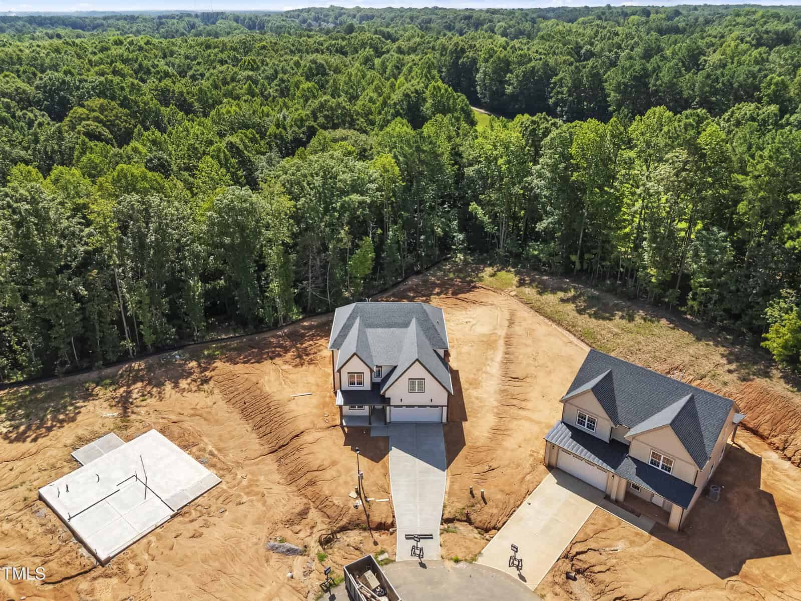 Aerial view of two newly built houses with gray roofs on large plots of cleared, bare earth, surrounded by dense green forest. A concrete foundation is visible on the left side. The driveway connects the homes to a paved road.