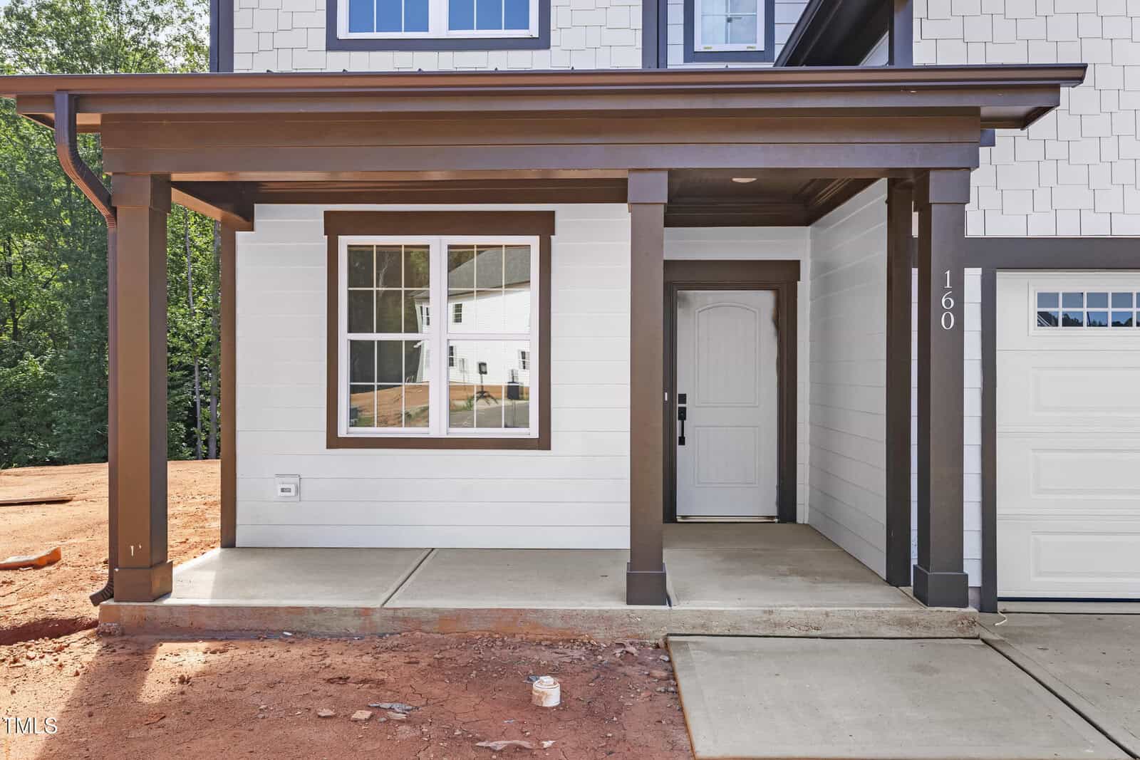A new house exterior with white siding, brown trim, a covered front porch with two pillars, a white door, a window, and a concrete walkway. The yard has bare red dirt and no landscaping yet.