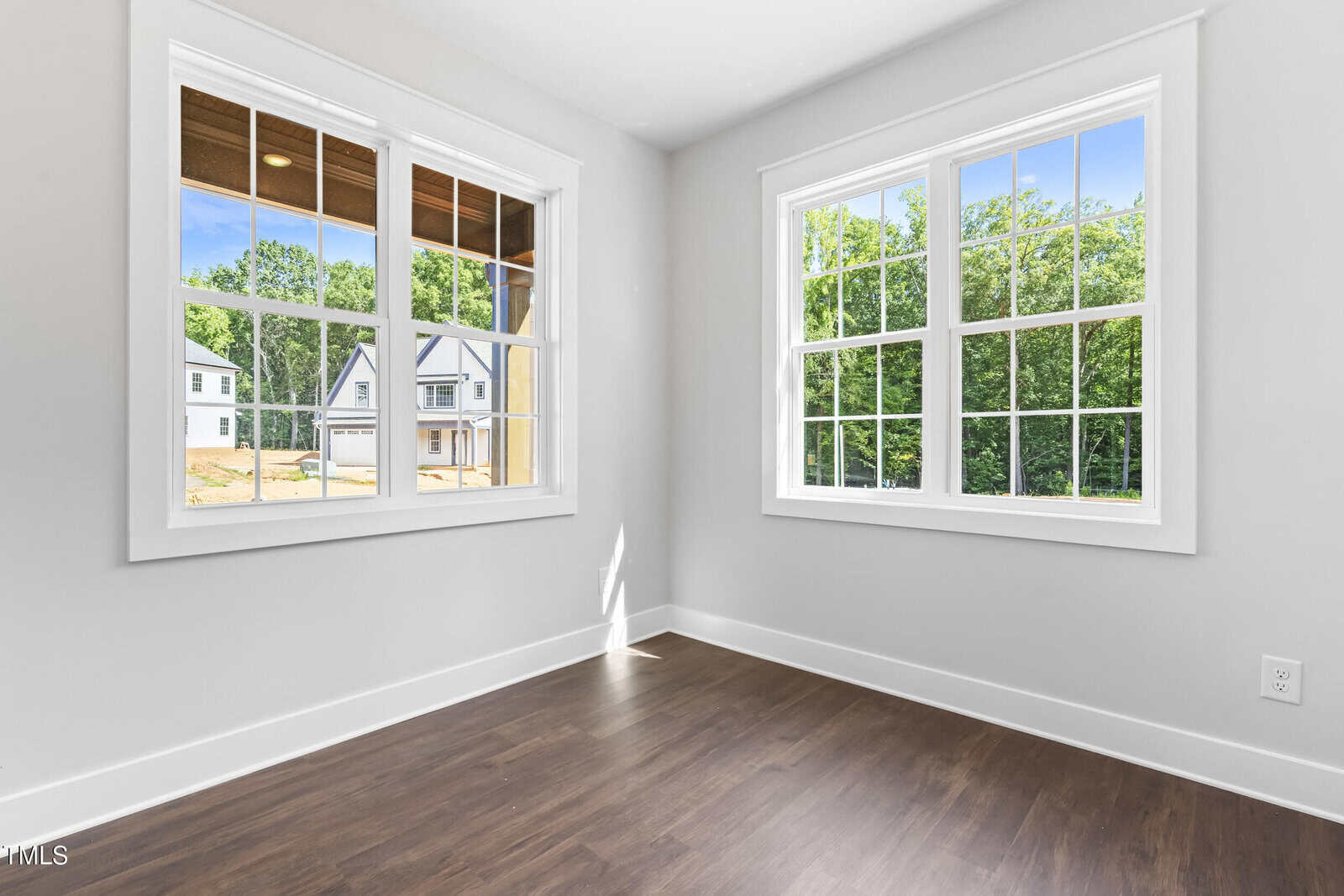 A bright, empty room with white walls, dark wood flooring, and two large windows showing a view of houses and trees outside on a sunny day.