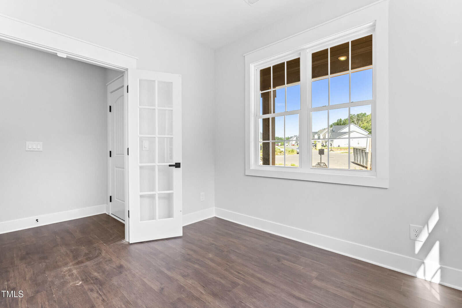 Bright room with white walls, dark wood flooring, a large window letting in natural light, and double French doors partially open to an adjacent space. Exterior view shows clear sky and houses.