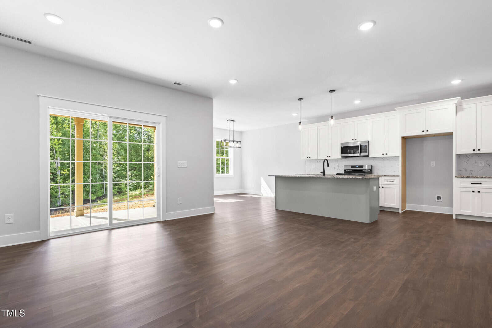 Spacious open-concept kitchen and living area with white cabinets, stainless steel appliances, large island, wood flooring, recessed lighting, and sliding glass doors leading to a patio with a view of greenery outside.