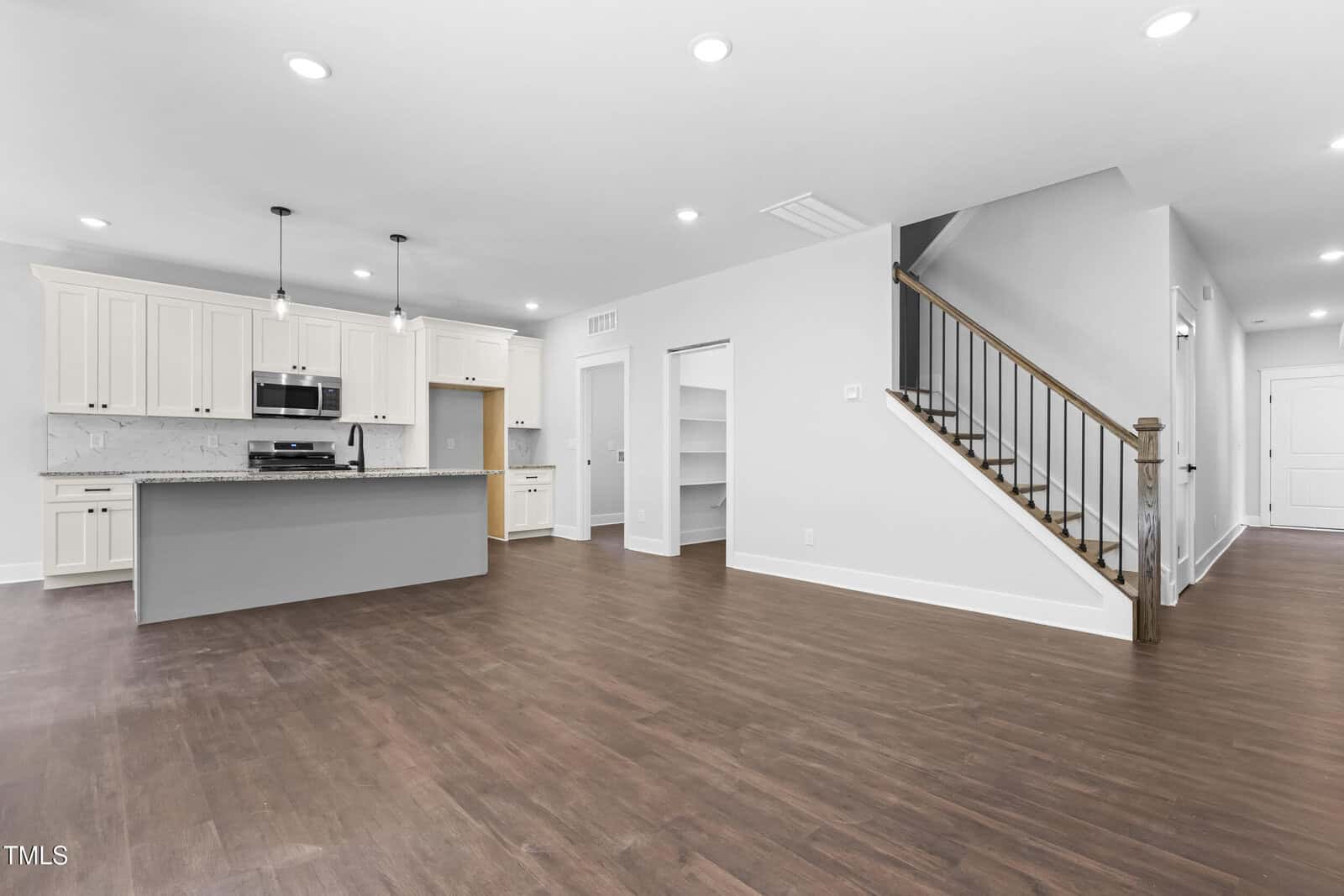 Open modern kitchen with white cabinets, stainless steel appliances, and a large island. Adjacent open area with wood flooring, visible staircase with wooden railing, and recessed lighting throughout the space.