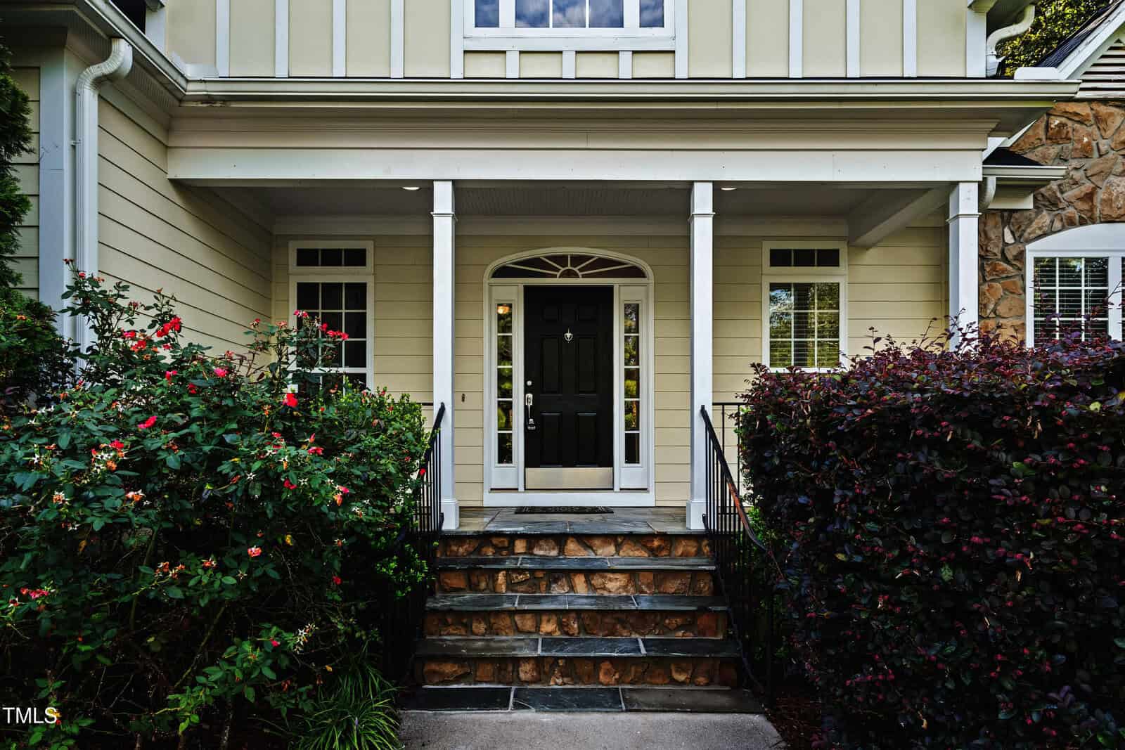 Front entrance of a house with a black door, stone steps, white trim, and beige siding, flanked by railings and lush bushes with red flowers and green leaves on both sides.
