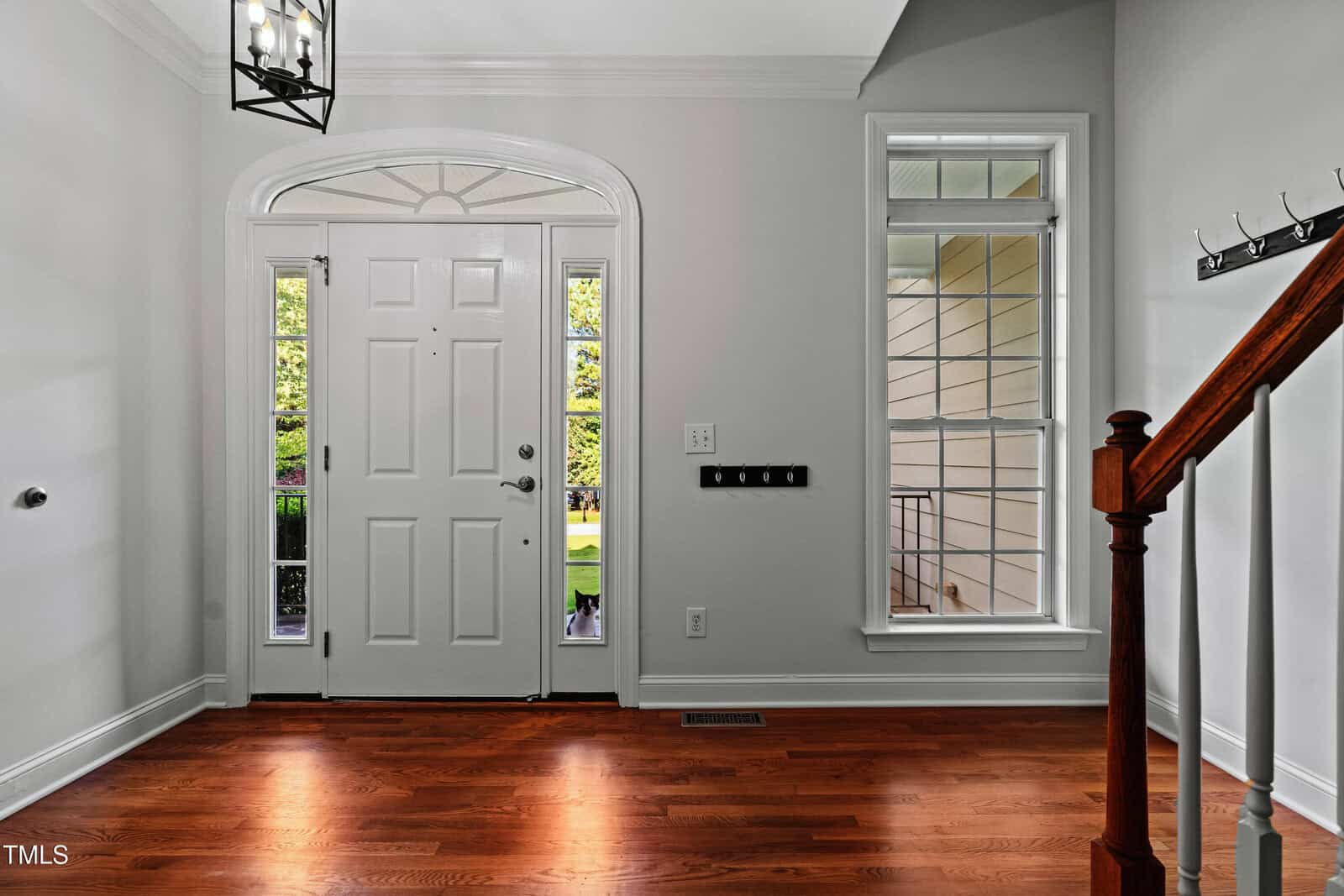 Bright entryway with a white front door, large side window, wooden floor, staircase with a wooden banister, light gray walls, and a modern light fixture overhead. Greenery is visible through the door’s window.
