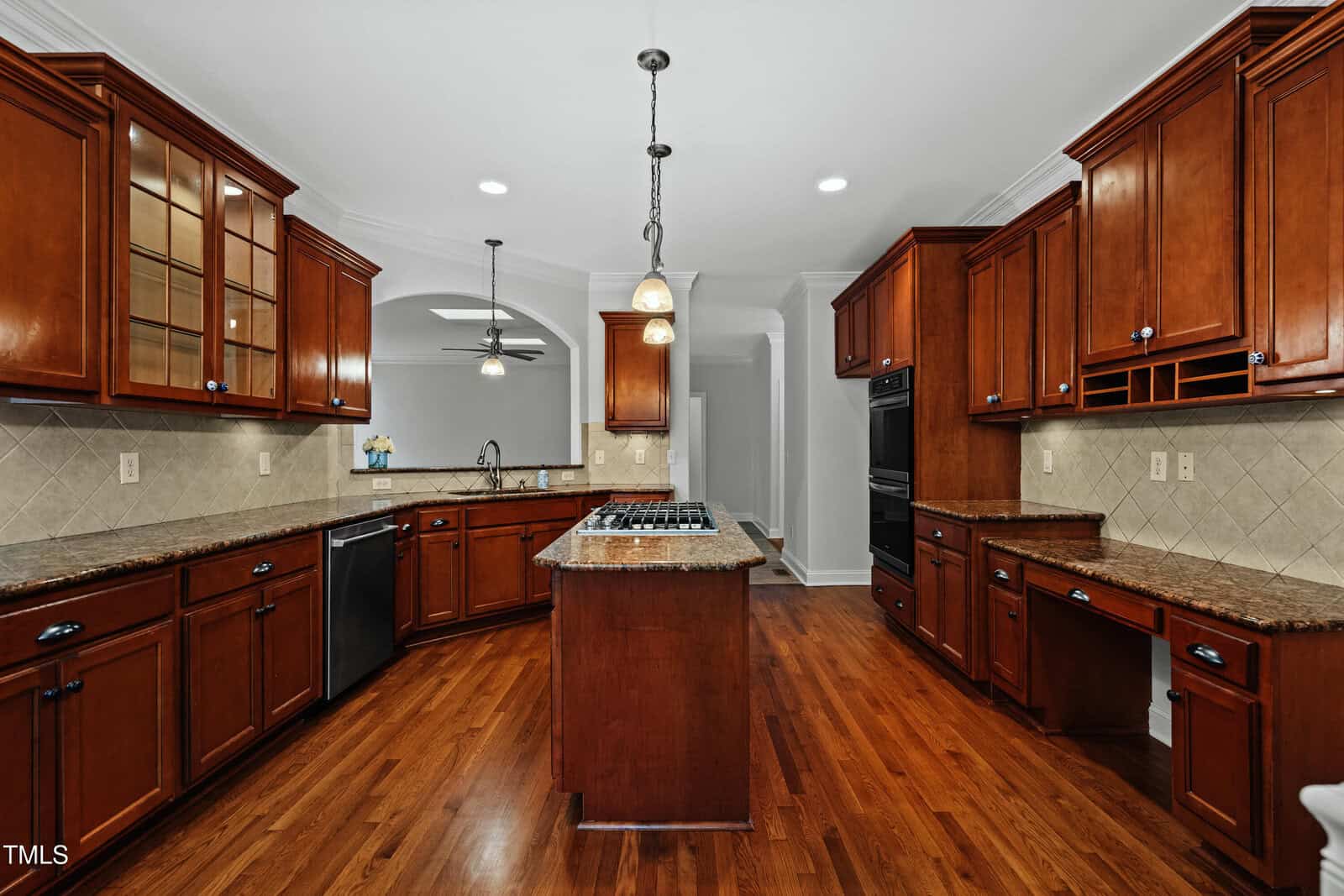 Spacious kitchen with wooden cabinets, granite countertops, a center island with a cooktop, stainless steel appliances, and hardwood floors under bright pendant lighting.
