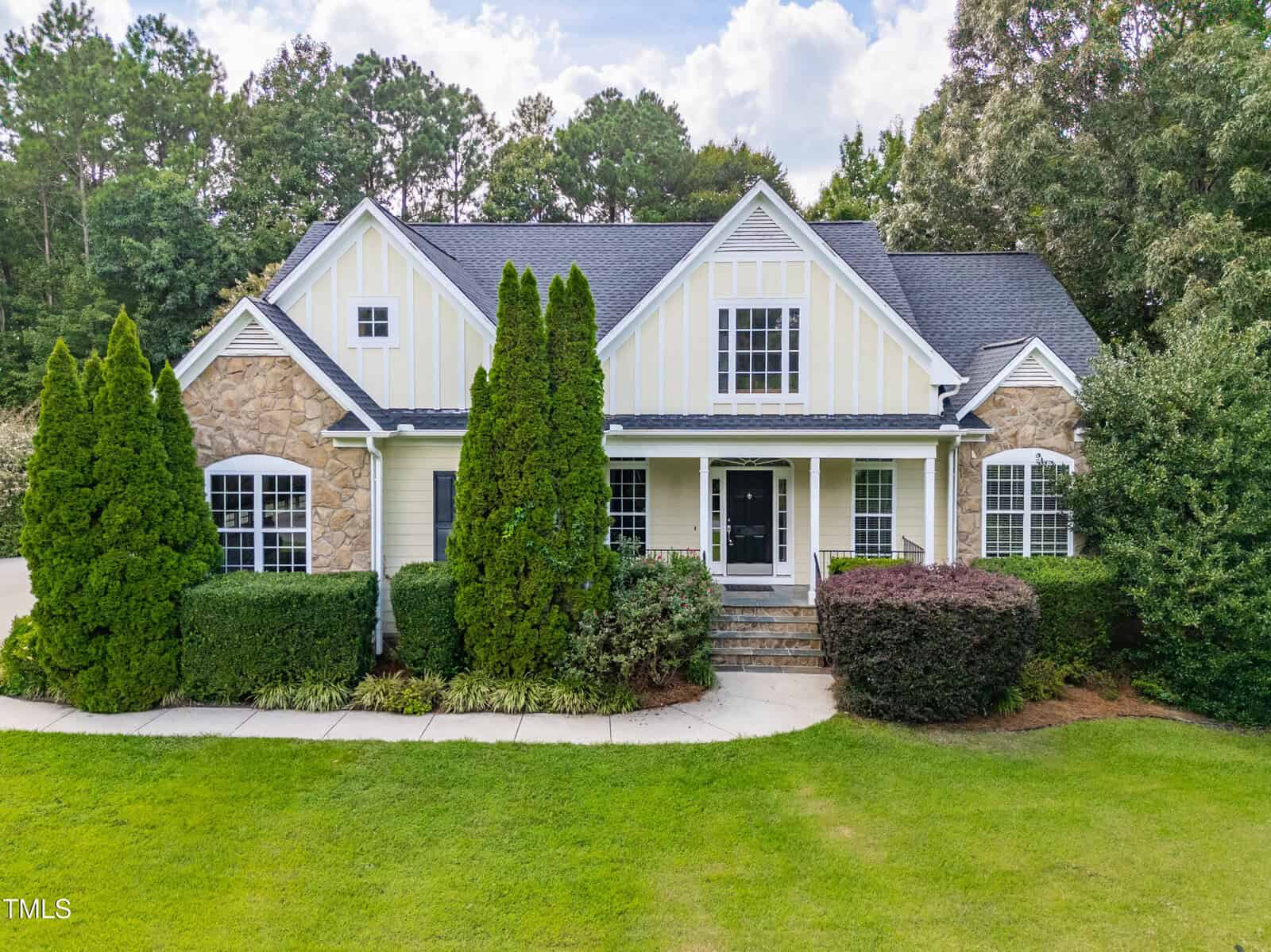 A two-story house at 509 Belmont Circle in Wake Forest features stone and yellow siding, a tall gabled roof, white trim, and a front porch. Tall shrubs and green bushes line the front, with a well-kept lawn and trees in the background.