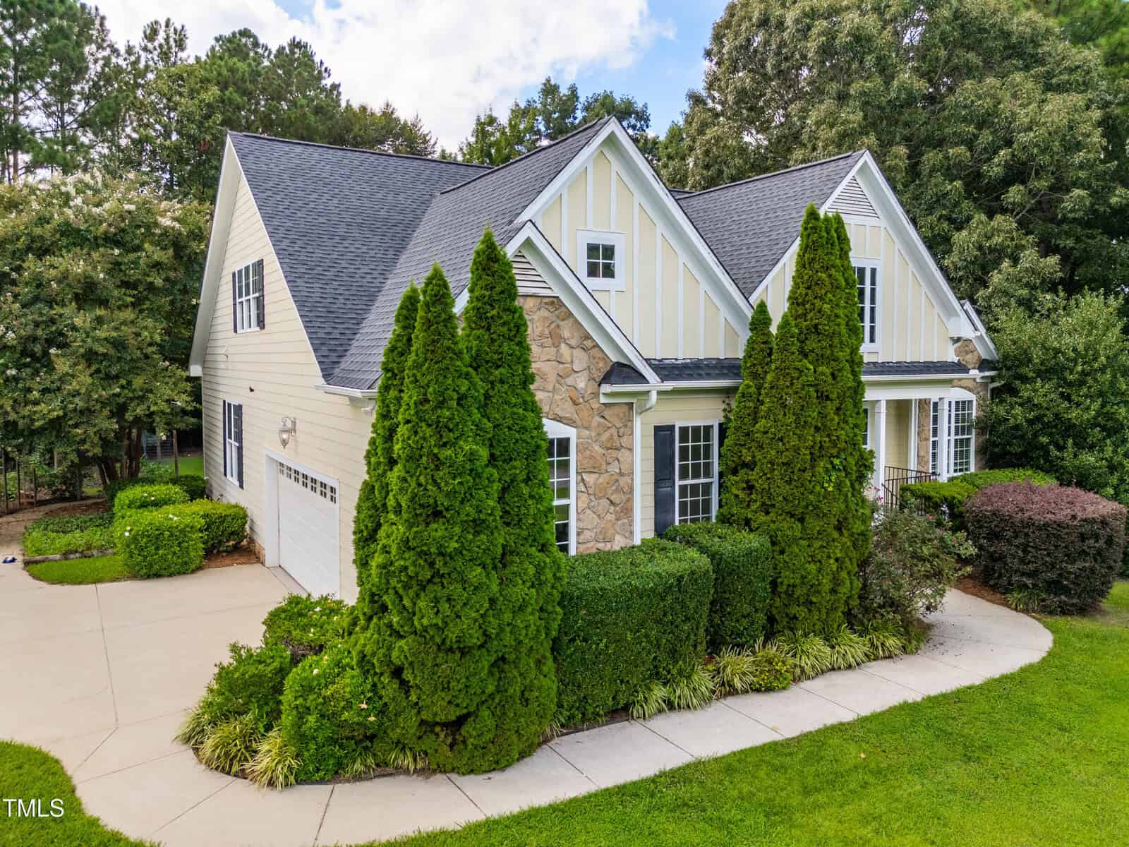 A two-story house with stone and light-colored siding, steep gable roofs, a double garage, tall evergreens in front, manicured bushes, and a concrete driveway surrounded by green lawn and trees.