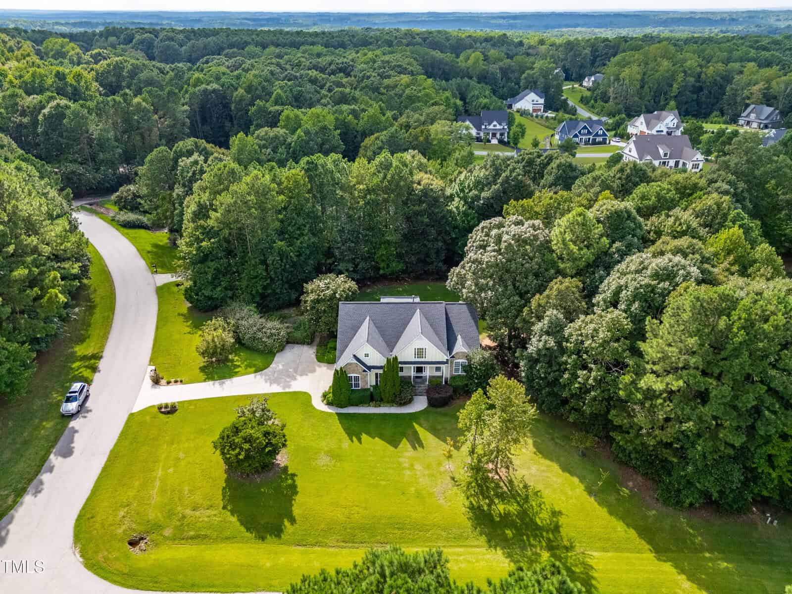 A large house with a white exterior and dark roof sits on a spacious, manicured lawn surrounded by trees, with a curved driveway and a quiet street in a wooded suburban neighborhood.