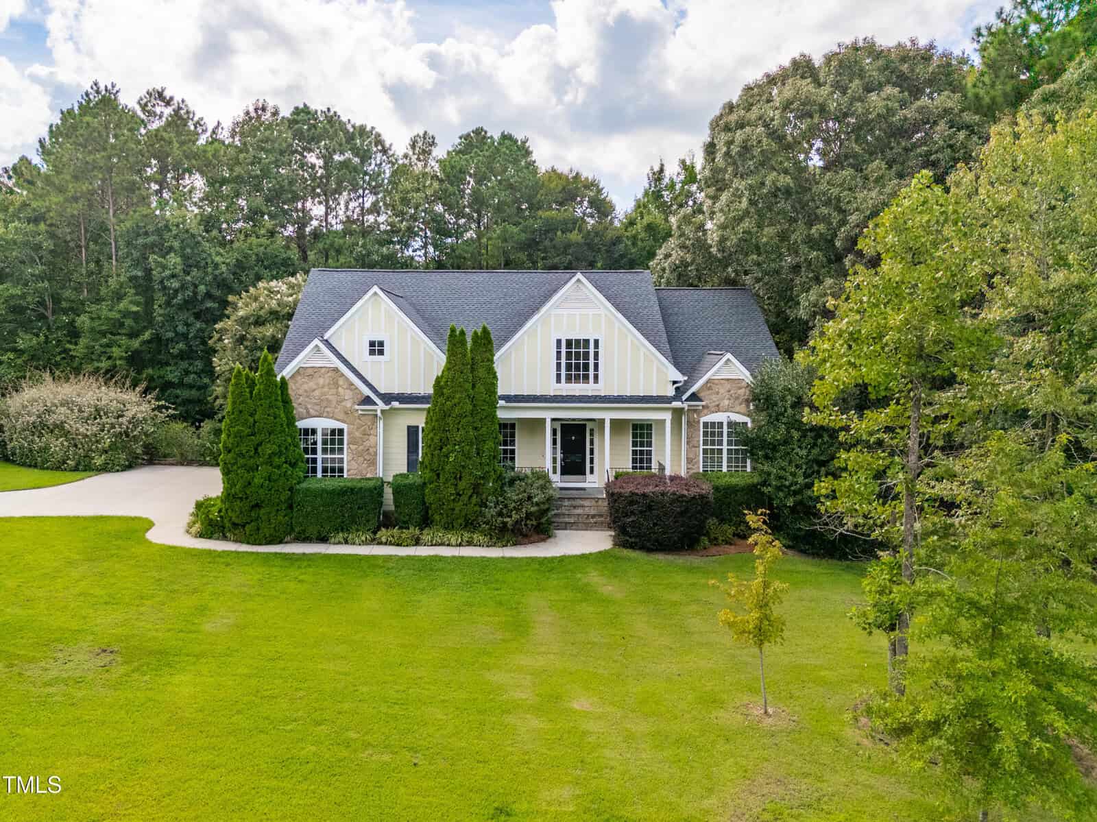 A two-story house with a gabled roof, yellow and white exterior, brick accents, and large front windows, surrounded by green lawn, tall trees, and a concrete driveway.