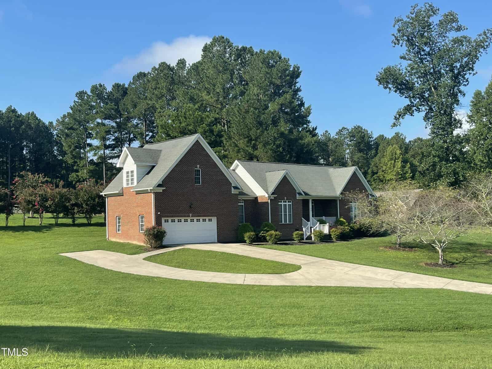 A red brick house with a gray roof and attached garage sits on a large, neatly mowed lawn. A curved driveway leads to the garage. The property is surrounded by trees under a clear blue sky.