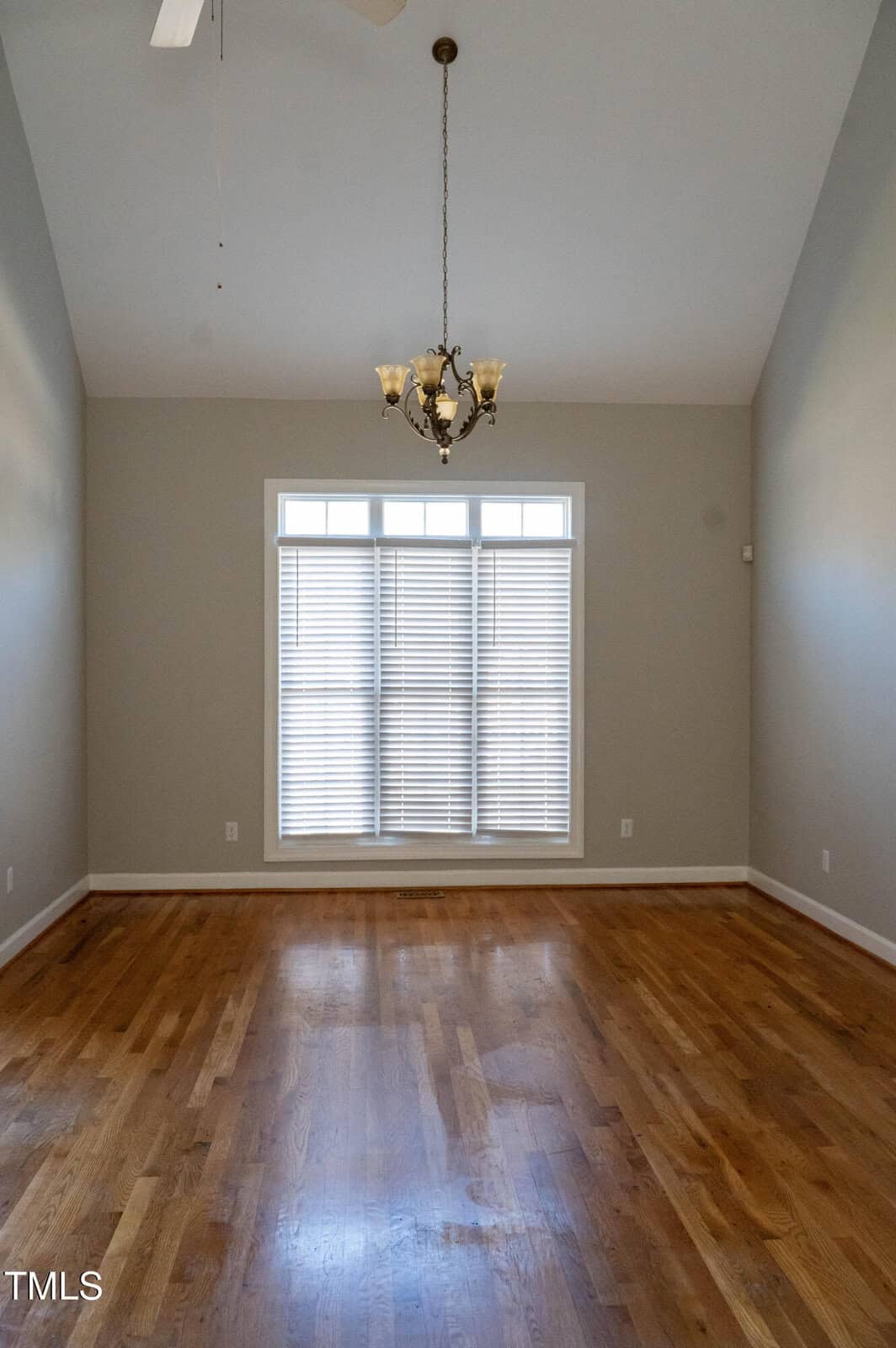 An empty room with light gray walls, a wooden floor, a chandelier, and a large window with white blinds allowing some natural light in. The ceiling is vaulted, and the room has no furniture.