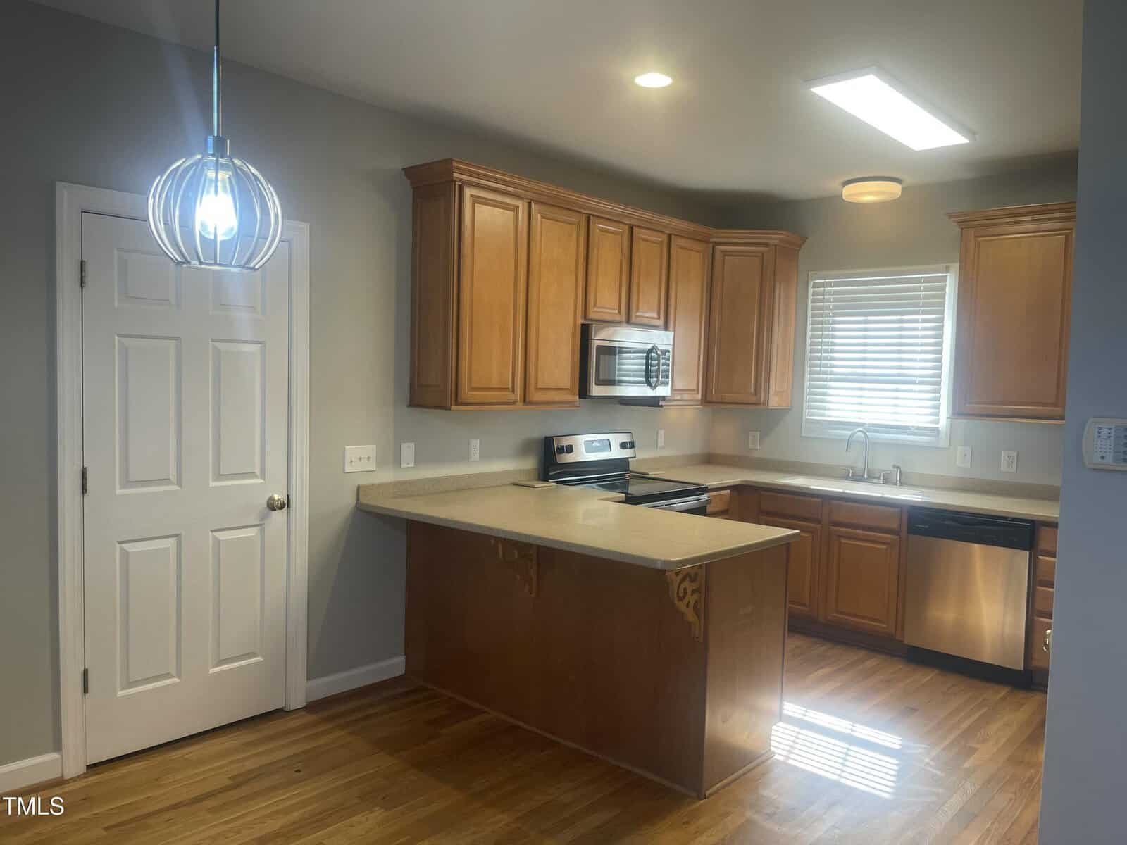 A kitchen with wooden cabinets, stainless steel appliances, a double sink below a window with blinds, a pendant light fixture, and a peninsula counter with corbels. Hardwood flooring and neutral walls are visible.