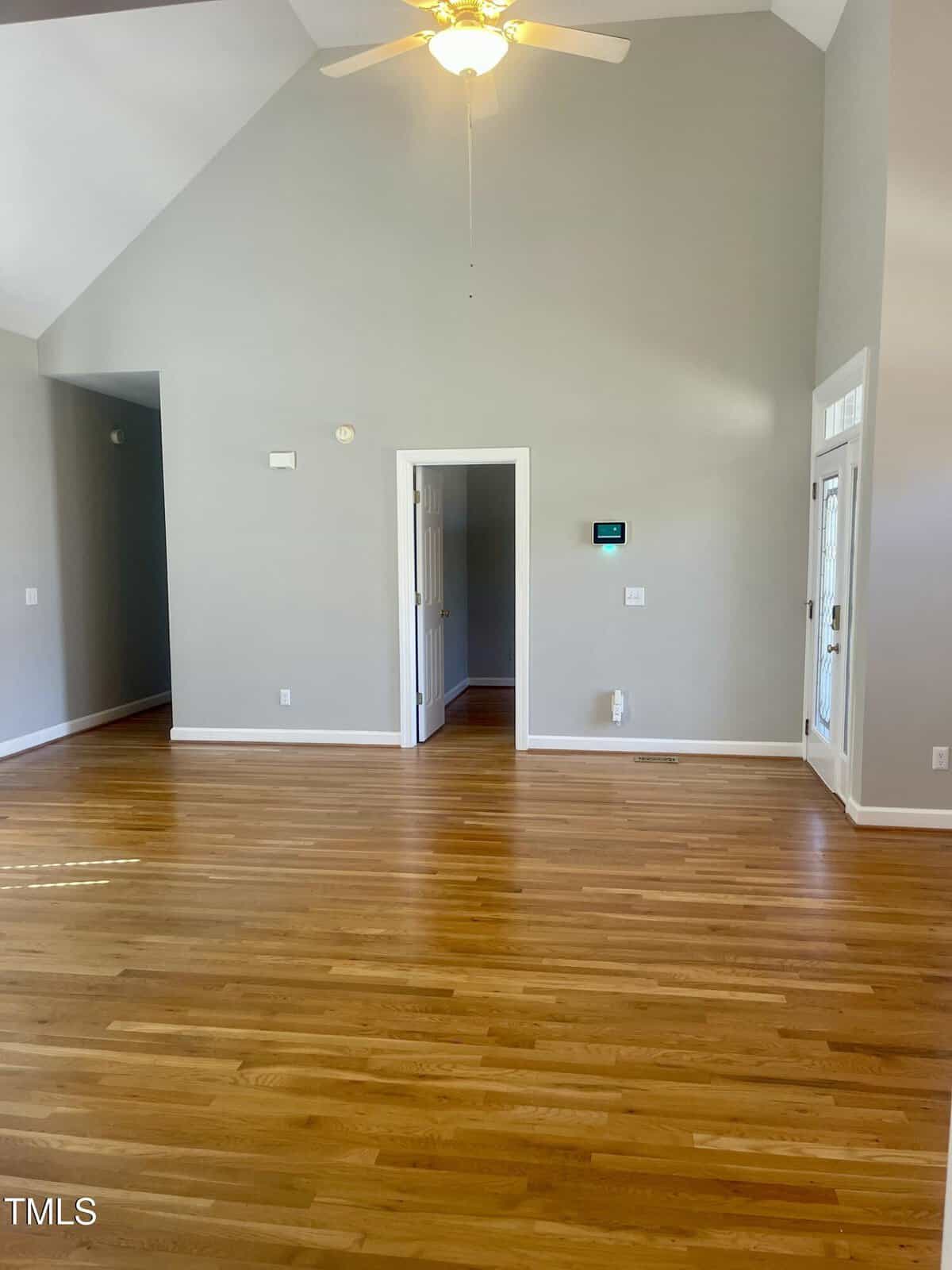 A spacious, empty room with light hardwood floors, gray walls, a ceiling fan with light, vaulted ceilings, and a door leading to another room. A thermostat and vent are visible on one wall.
