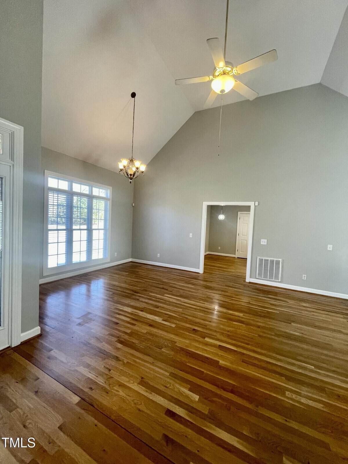 Spacious, empty living room with high vaulted ceiling, gray walls, hardwood floors, large windows, ceiling fan, and chandelier. Doorway leads to another room with a light fixture.