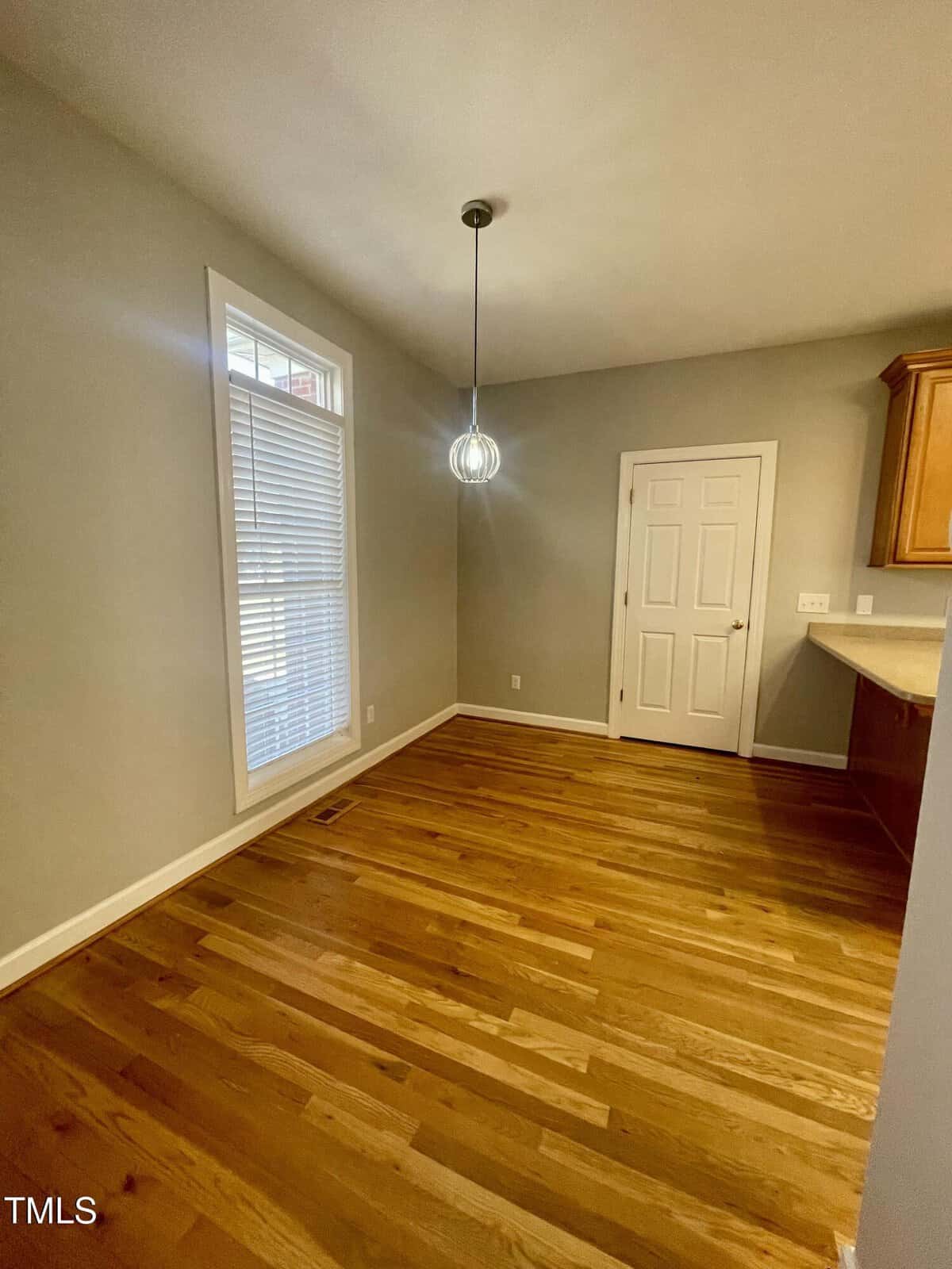 A small dining area with hardwood floors, a large window with blinds, a modern pendant light, beige walls, and a white door next to a kitchen counter with wooden cabinets.