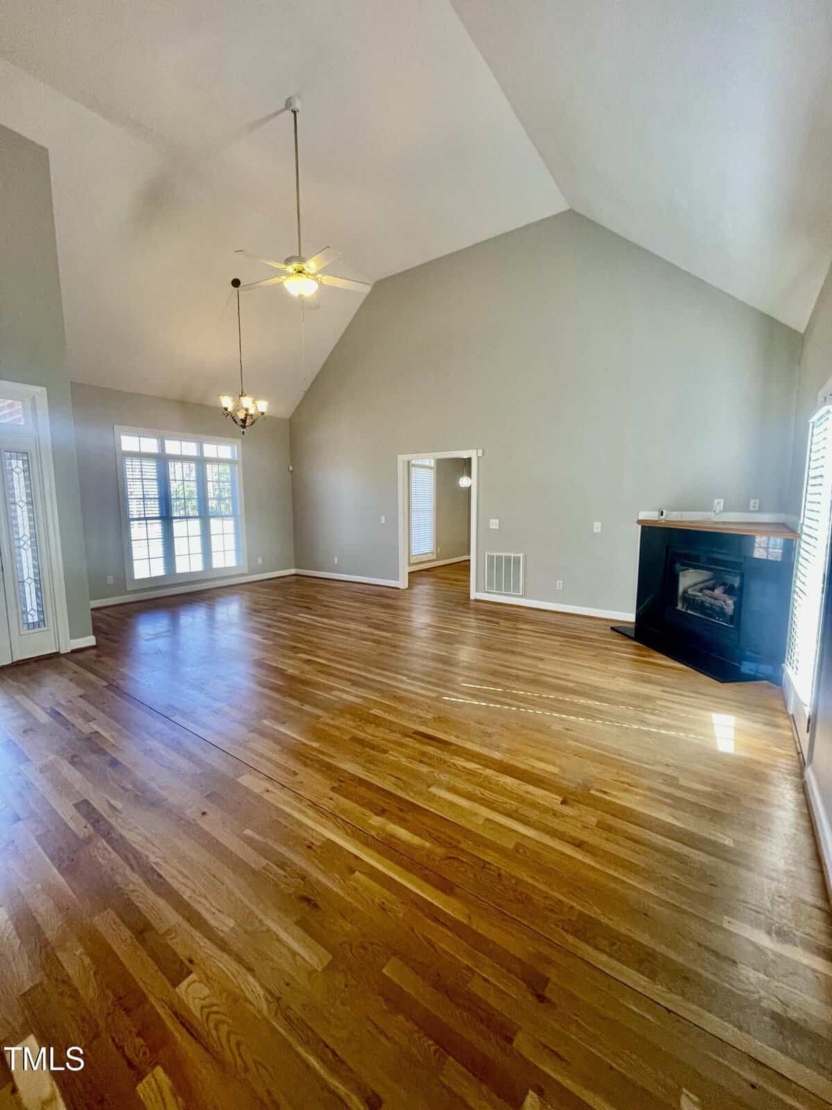 Spacious living room with vaulted ceilings, hardwood floors, large windows, two ceiling fans, and a corner fireplace. Sunlight streams in through the windows, brightening the open, empty space.