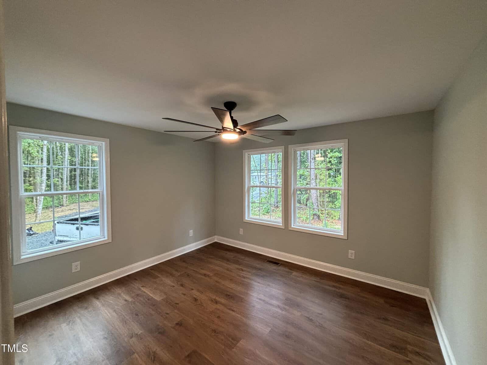 Empty room with light gray walls, two large windows, a ceiling fan with light, and wood flooring. Trees and greenery are visible outside the windows, and natural light brightens the space.