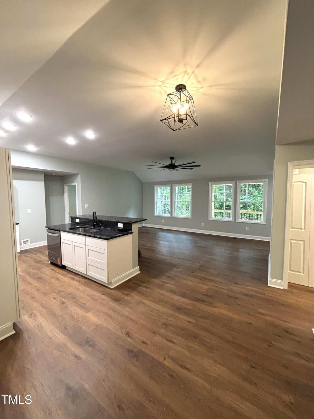 Open-concept living space with wood flooring, a kitchen island with a dark countertop, modern ceiling lights, large windows, and a ceiling fan. Neutral walls and white trim complete the interior.