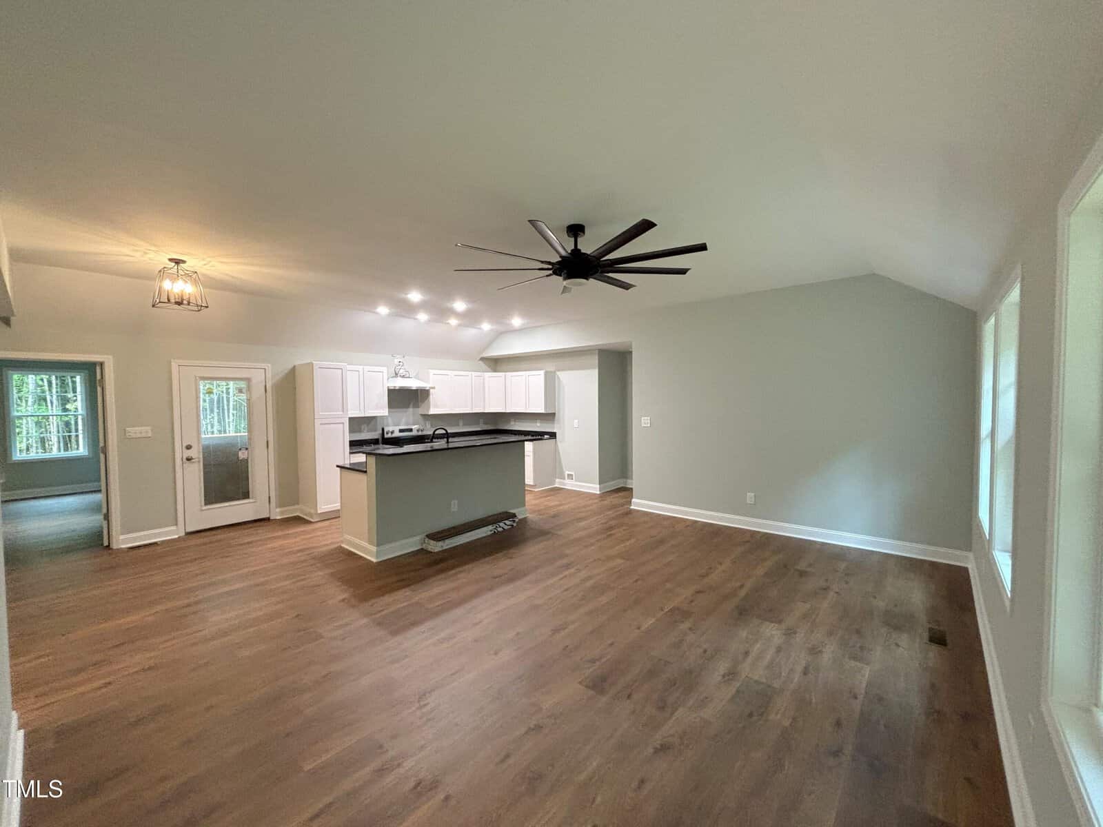 A spacious open-concept room with wood flooring, white kitchen cabinets, a large black ceiling fan, multiple ceiling lights, and several windows letting in natural light. A kitchen island separates the kitchen from the living area.