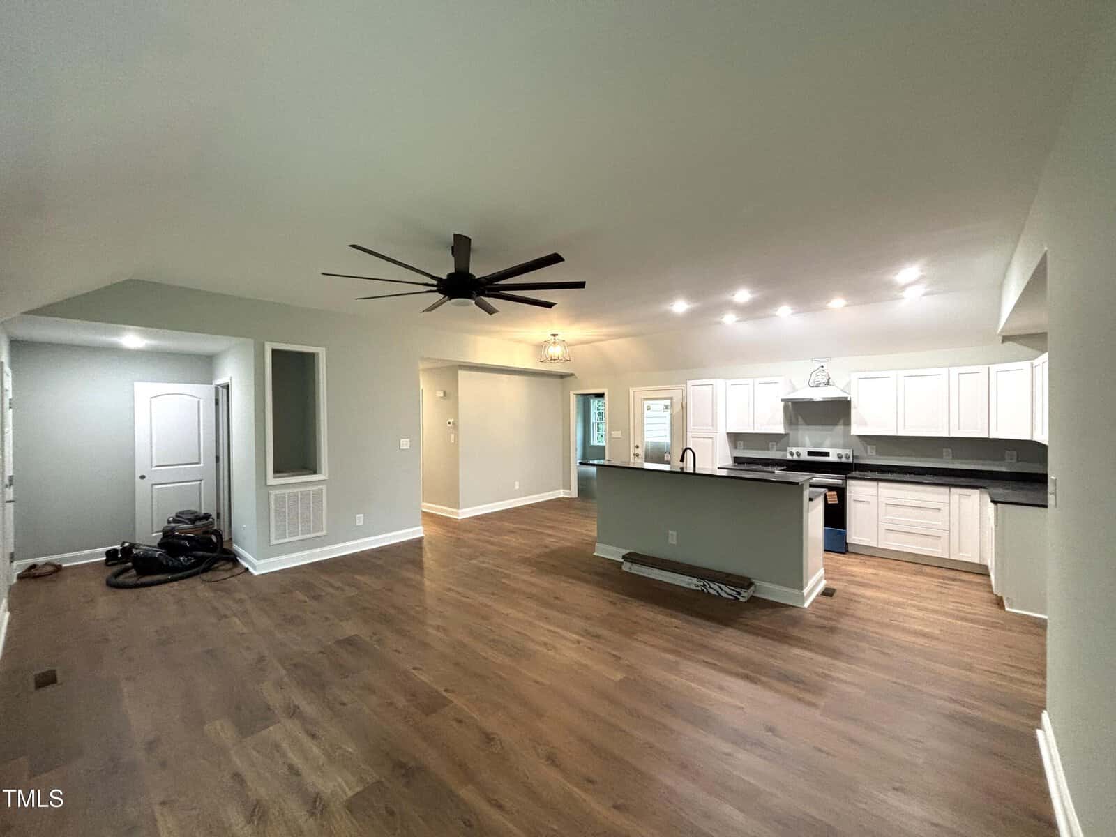 Open-concept living area with wood flooring, ceiling fan, and recessed lighting. The modern kitchen features white cabinets, stainless steel appliances, and a large island. Some tools and equipment are on the floor near the entryway.
