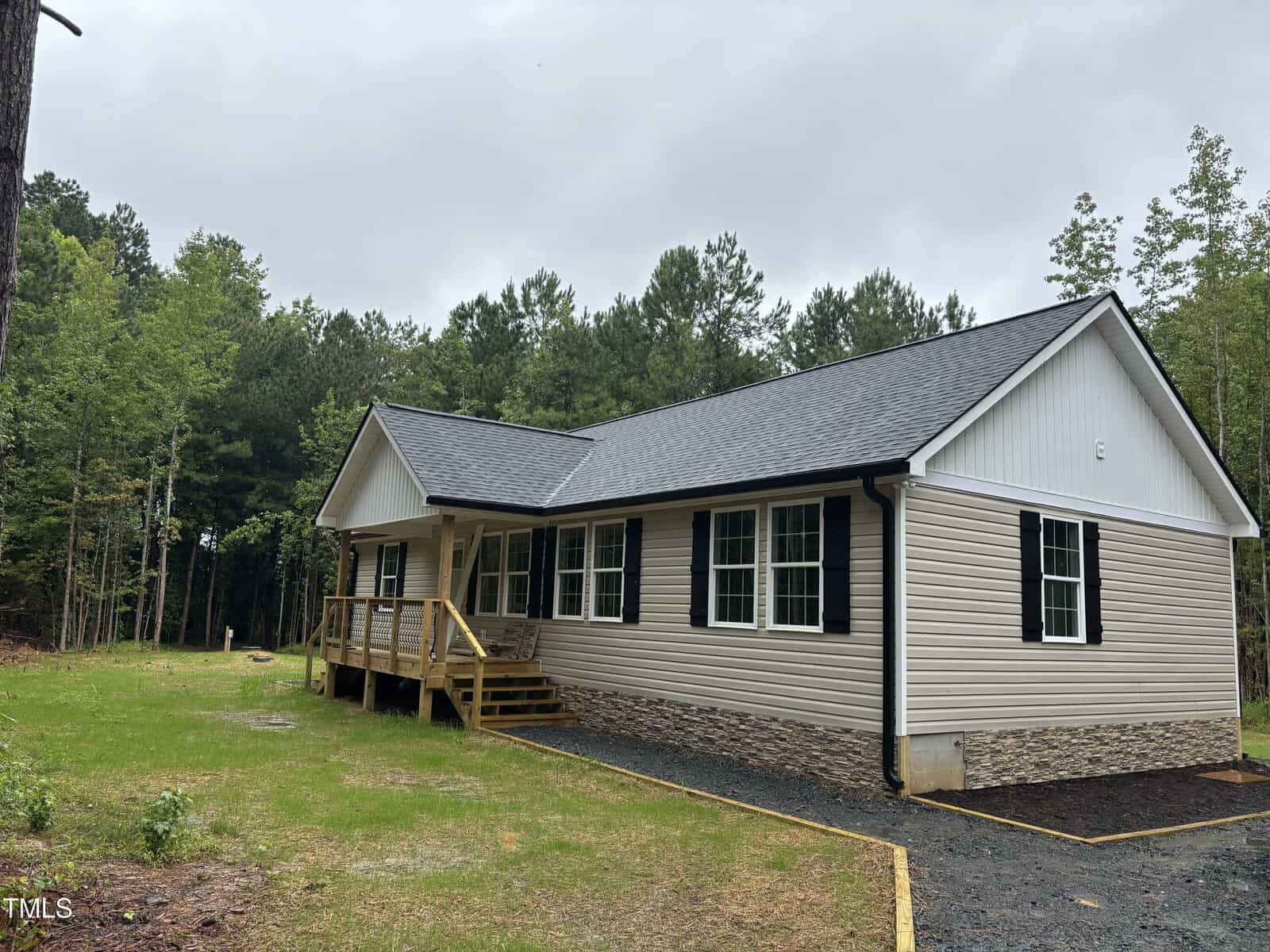 A single-story beige house with black shutters and a dark gray roof sits on a grassy lot bordered by trees. A small covered porch with wooden steps leads to the front door. The sky is overcast.