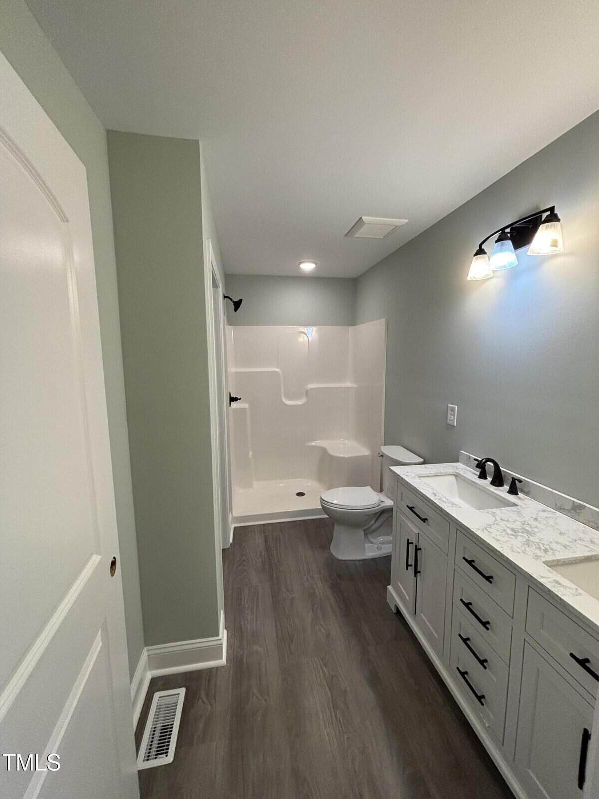 Modern bathroom with light green walls, wood-look flooring, a white vanity with marble countertop, a toilet, and a white shower. Black light fixture above the vanity. Vent on the floor near the door.