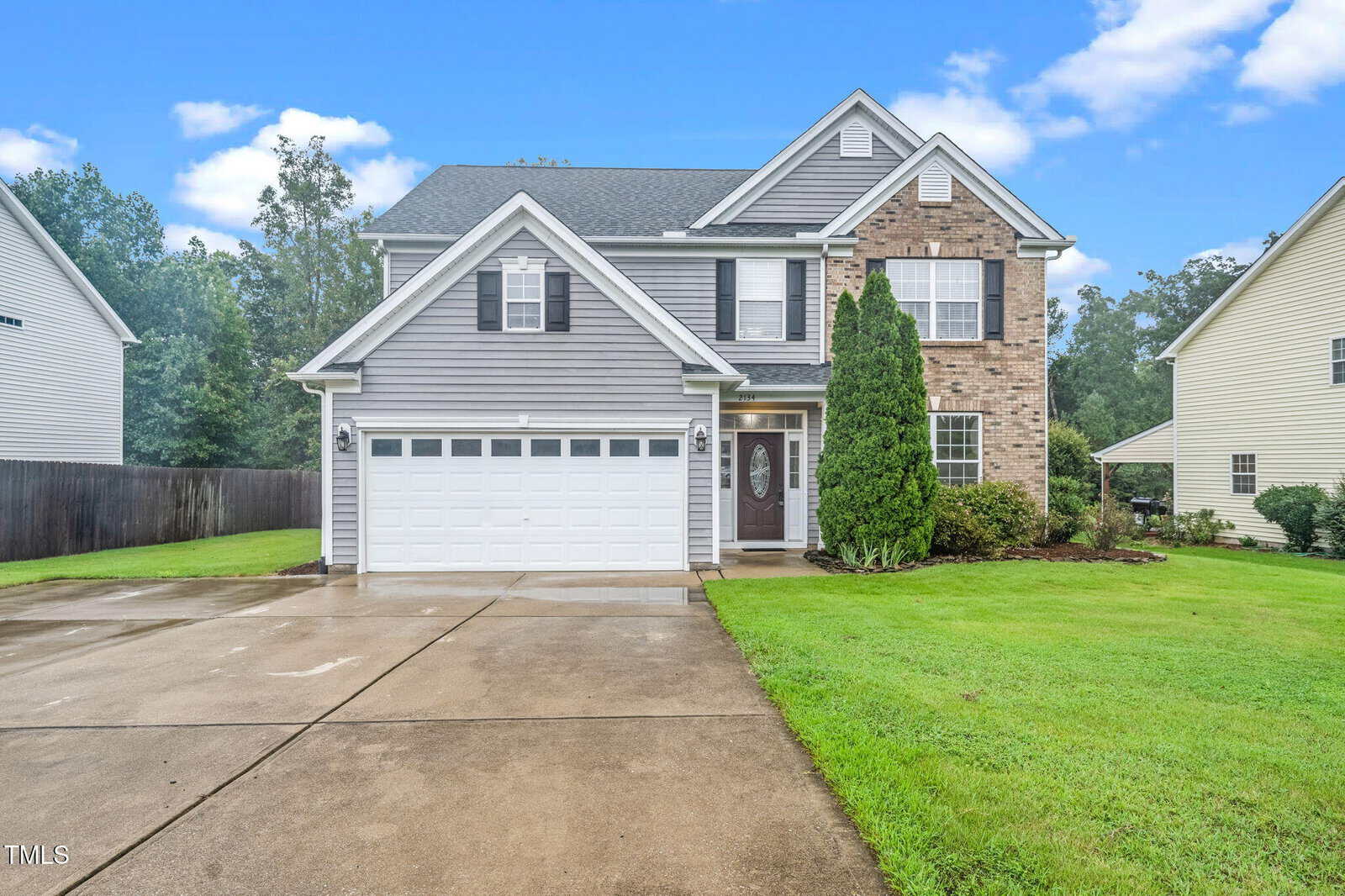 A two-story suburban house at 2134 Westbourne Drive, Creedmoor, features gray siding, brick accents, a white double garage door, and a well-kept lawn, pictured on a cloudy day with a wet driveway.