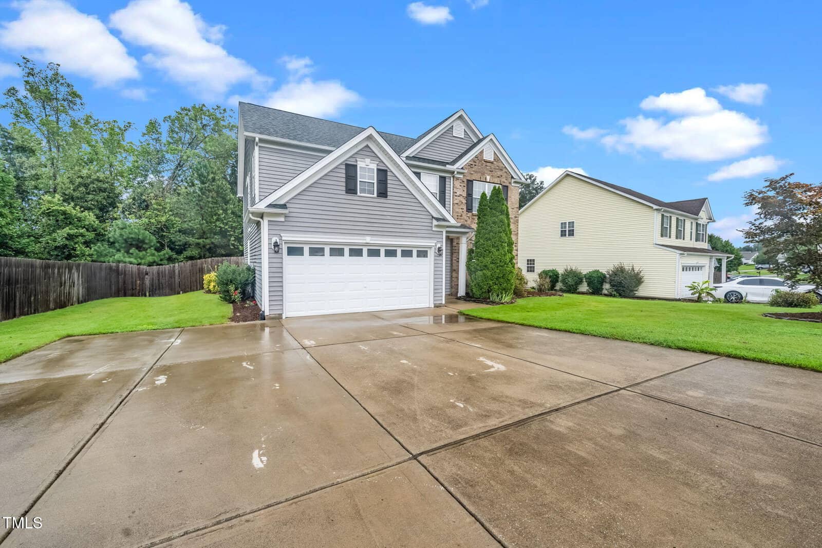 A two-story suburban house with gray siding, white trim, and a double garage is shown. The concrete driveway is wet, and the lawn and trees are green under a blue sky with scattered clouds.