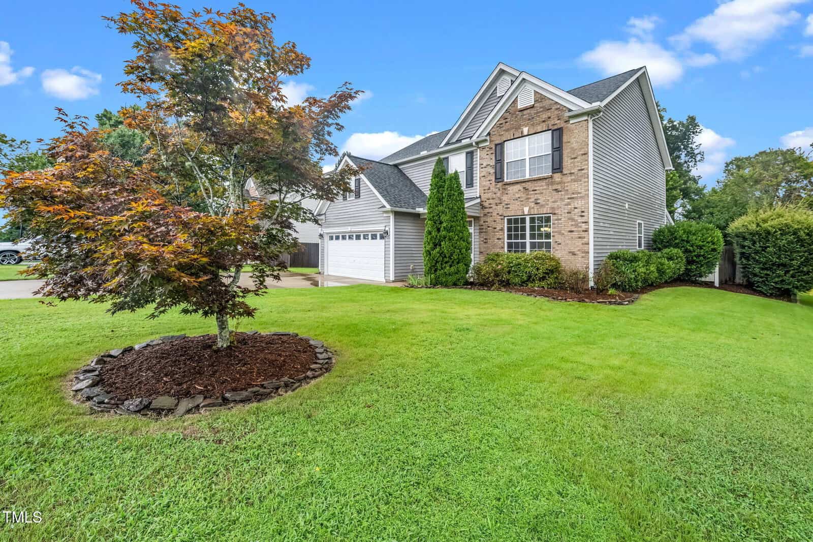 Two-story suburban house with brick and siding exterior, attached two-car garage, manicured lawn, landscaped bushes, and a small tree in the front yard under a partly cloudy sky.