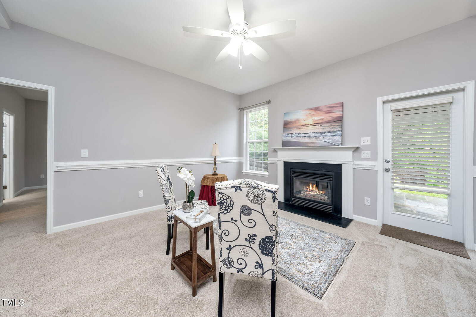 A small dining area with two patterned chairs and a table on beige carpet is set near a gas fireplace. A window and glass door let in natural light. The walls are light gray with white trim and a ceiling fan above.