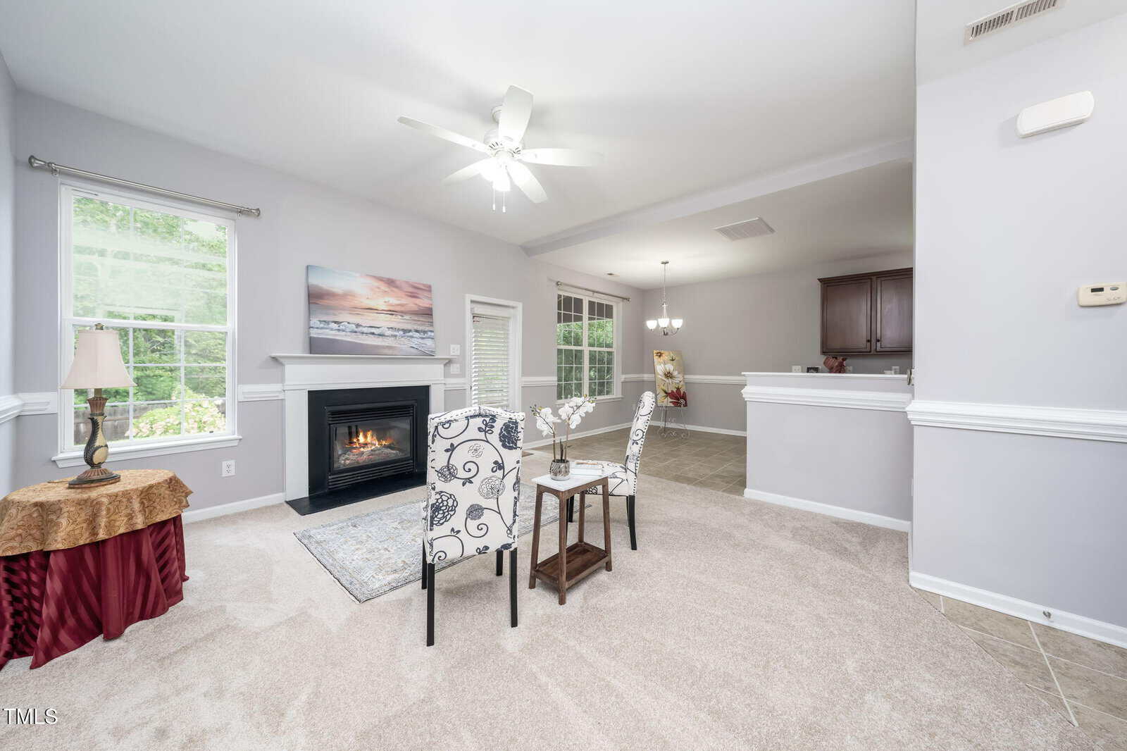 A living room with light carpet, a fireplace, two patterned chairs around a small table, and a window letting in natural light; an open dining area and kitchen are visible in the background.