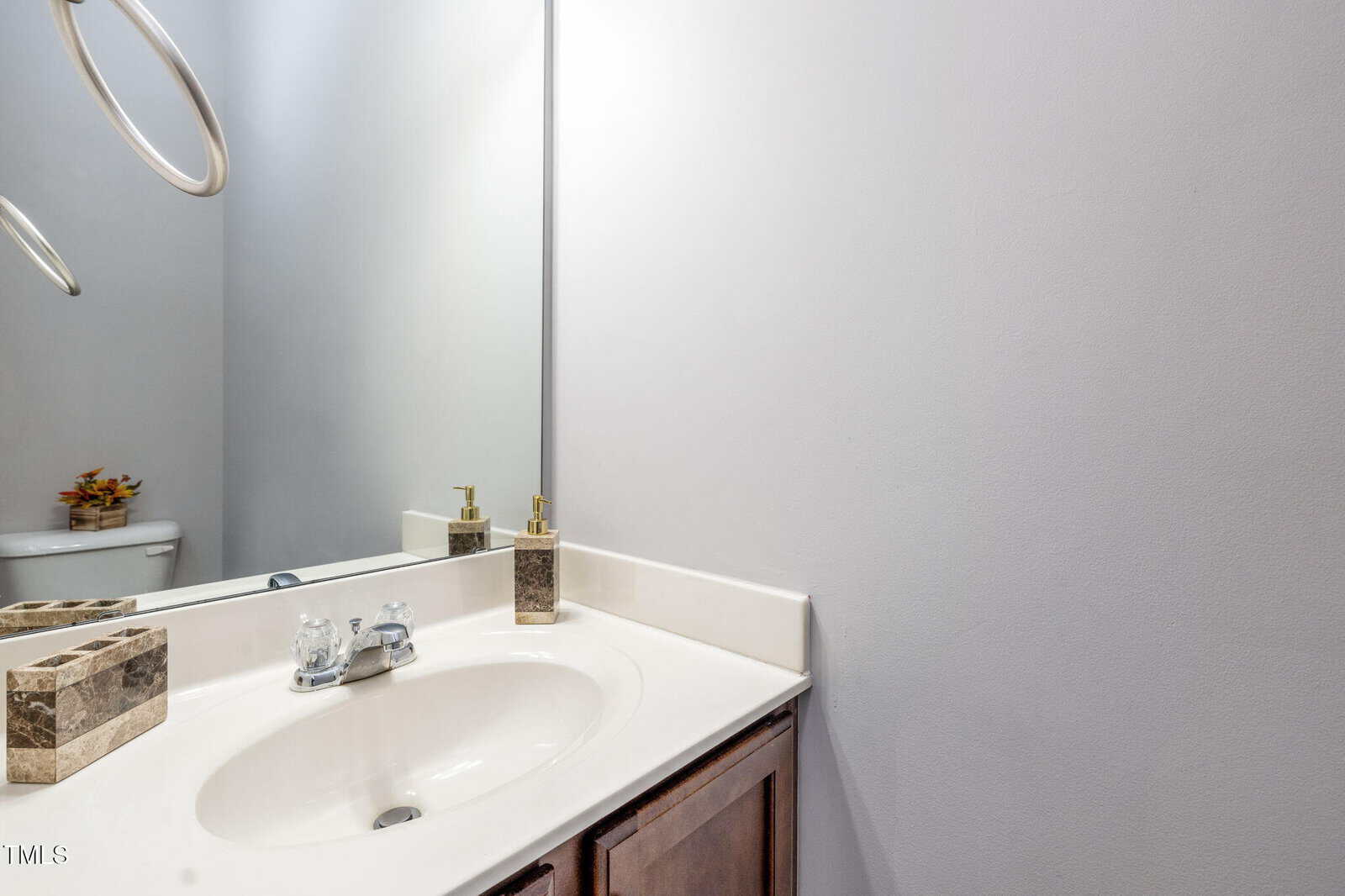 A bathroom with a white sink, chrome faucet, soap dispenser, and toothbrush holder on a beige countertop. A large mirror reflects part of the sink and a toilet with a small plant decoration.