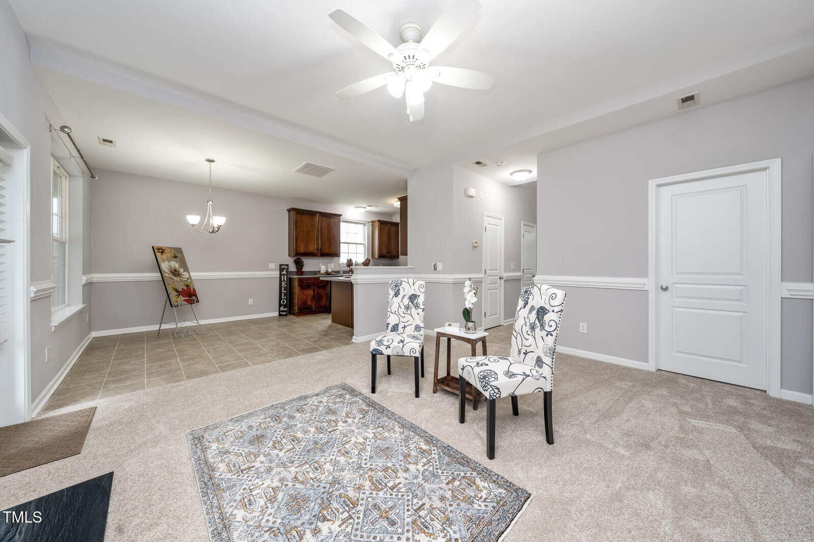A bright, open living space with light gray walls, beige carpet, a patterned area rug, two white and blue accent chairs, and a view into a tiled dining area and a kitchen with dark wood cabinets.