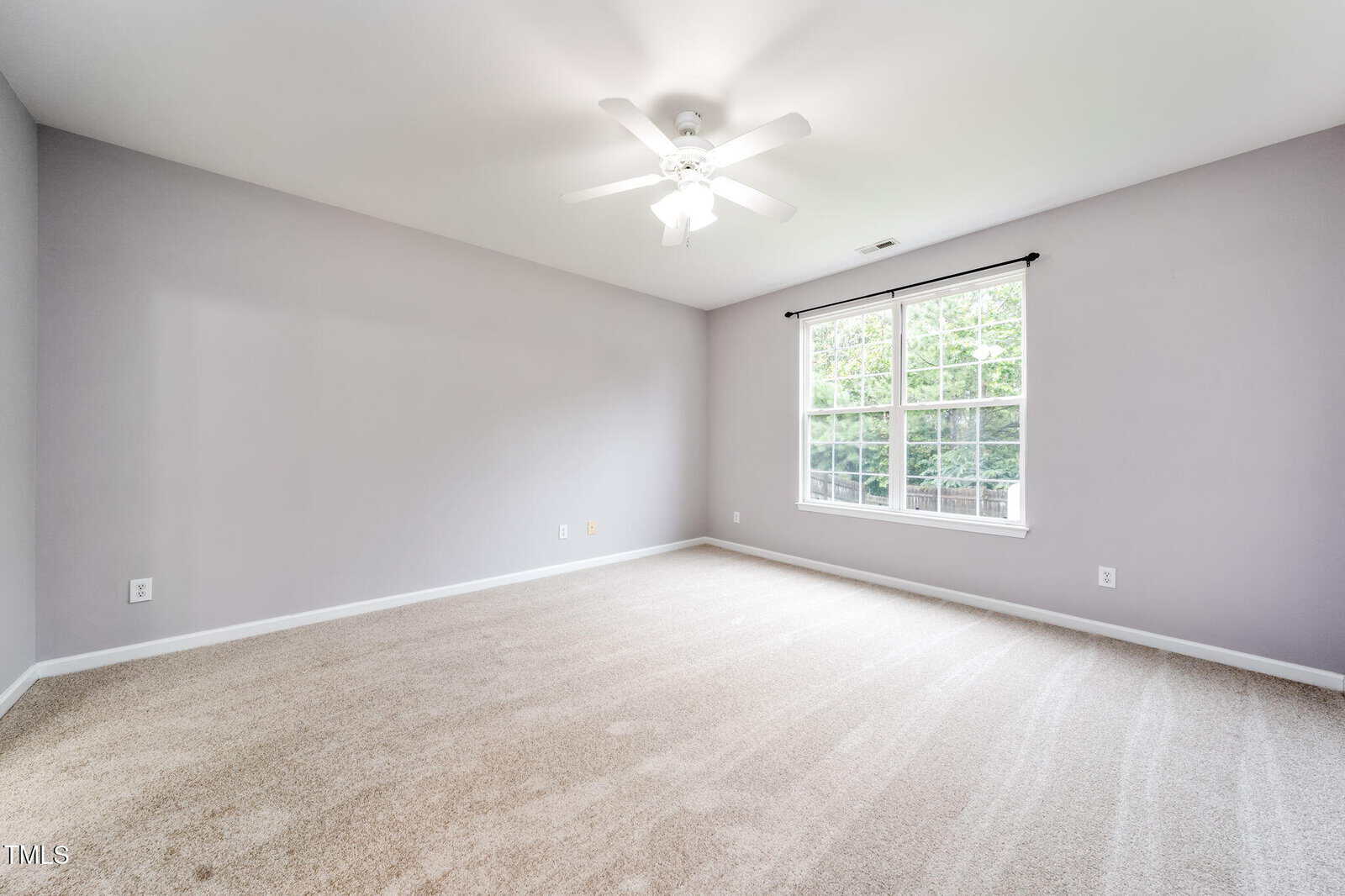 A spacious empty room with light beige carpet, light gray walls, a white ceiling fan, and a large window letting in natural light. The window provides a view of greenery outside.
