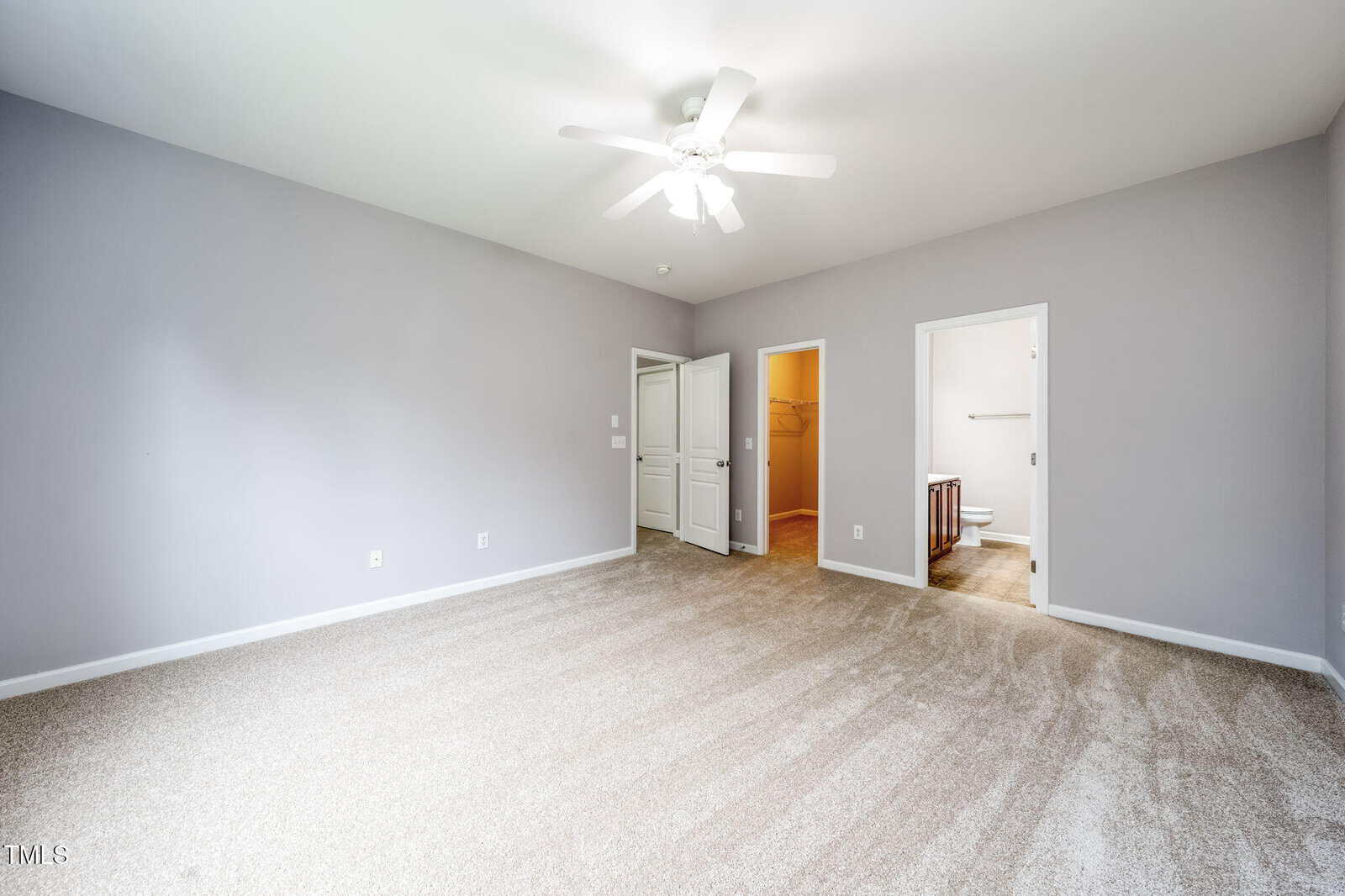 A spacious, empty bedroom with light gray walls, beige carpet, a white ceiling fan, and two open doorways leading to a walk-in closet and a bathroom. A closed white door is also visible on one wall.