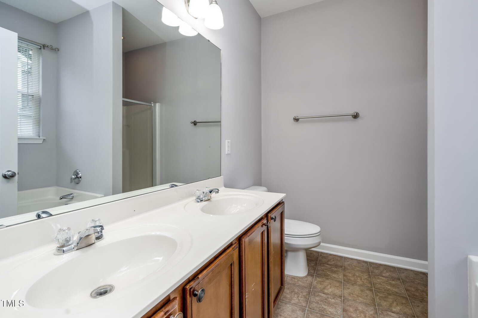 A bathroom with a double-sink vanity, large mirror, wooden cabinets, a toilet, towel bar, and a bathtub with a shower curtain, featuring gray walls and tile flooring.