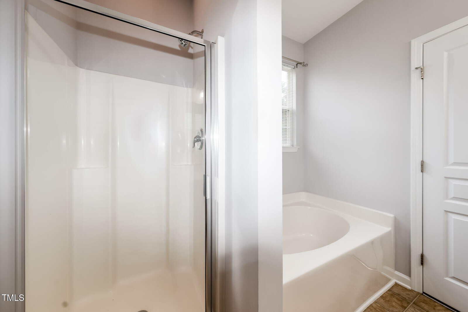 A bathroom with a glass shower on the left, a white bathtub on the right, a window above the tub, and a white door on the far right. The walls are painted light gray and the floor is tiled.