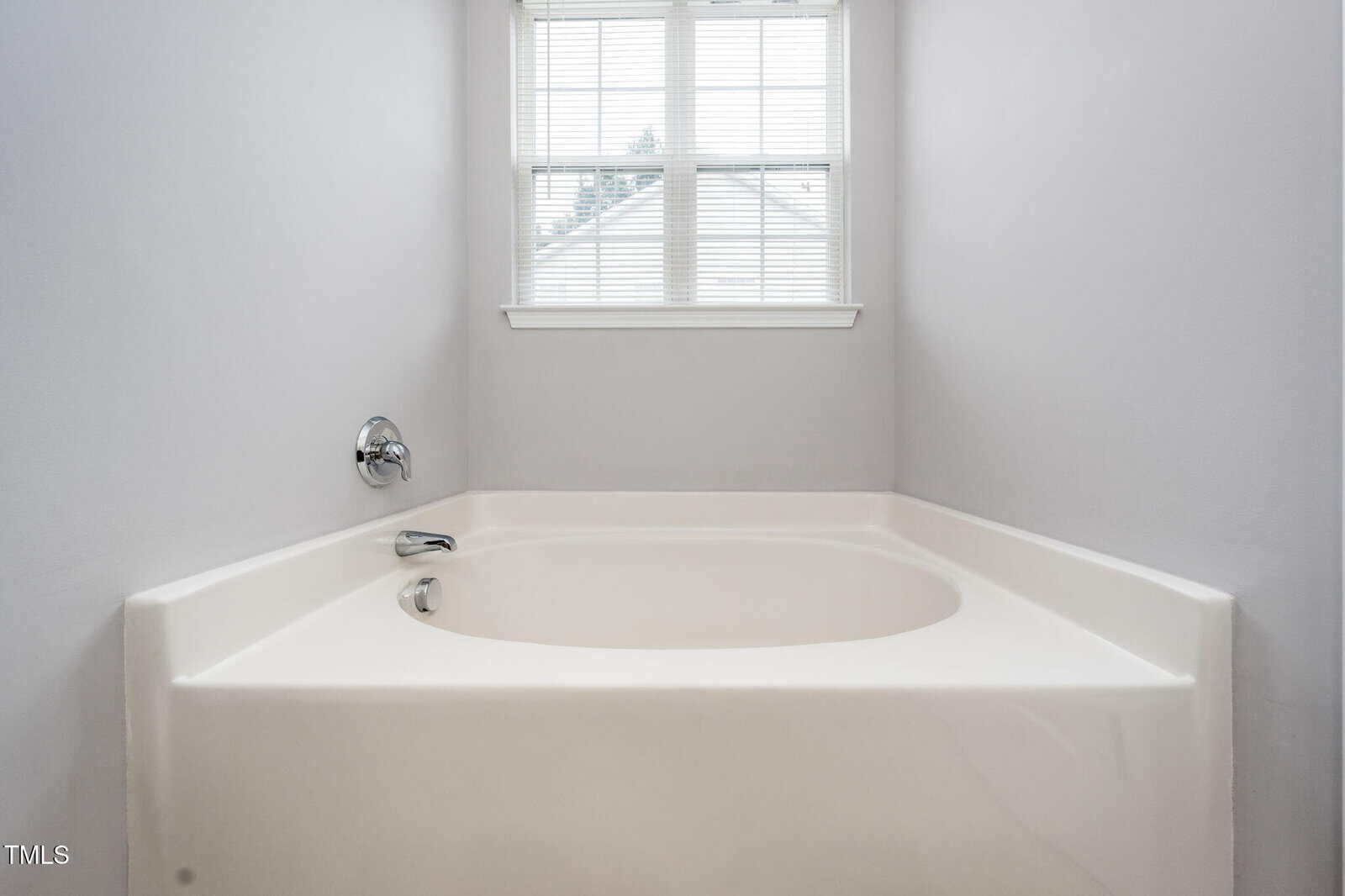 A white built-in bathtub sits beneath a window with closed blinds in a minimalist, light gray bathroom.