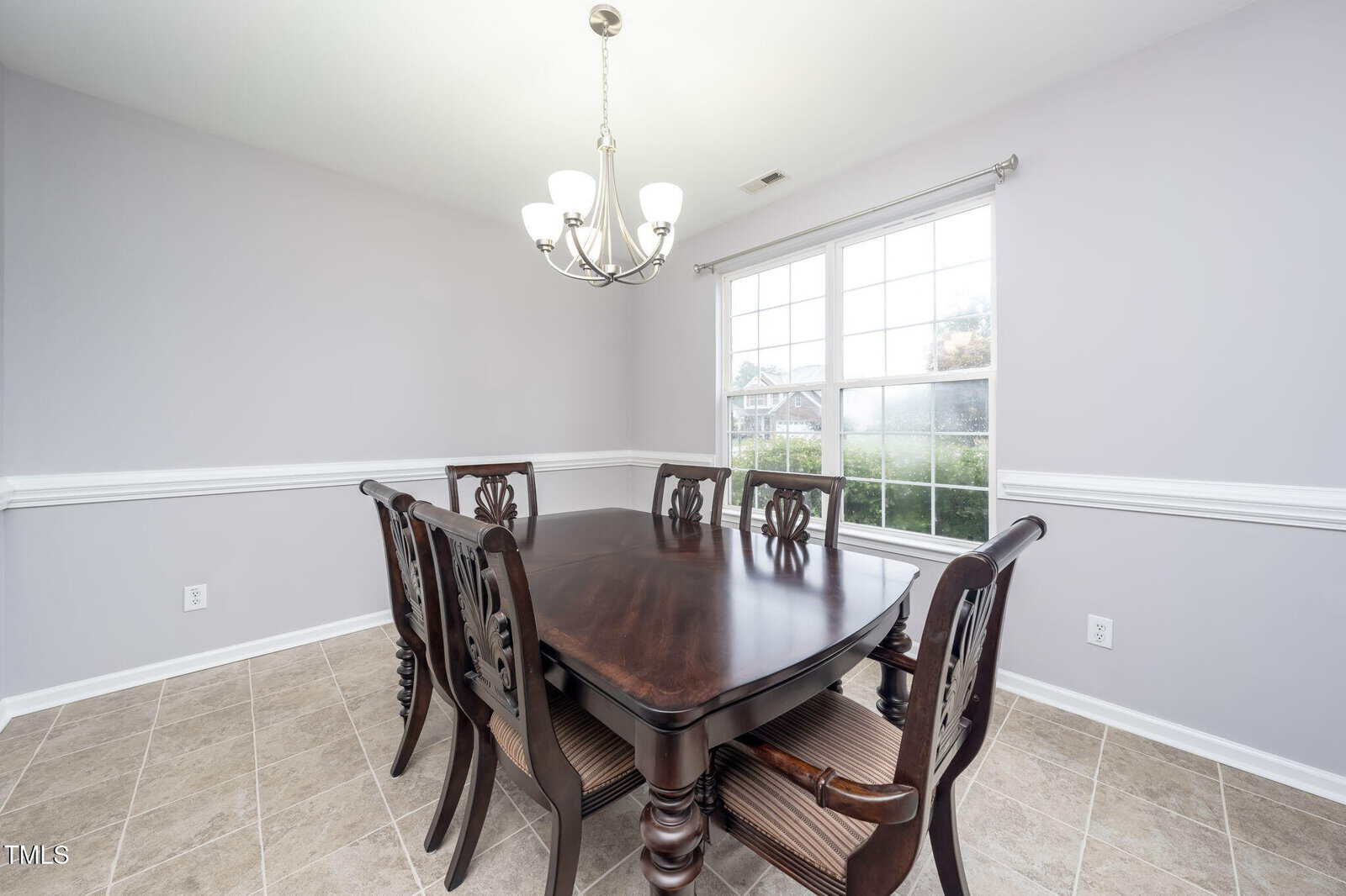 A dining room with a dark wooden table and six matching chairs, a modern chandelier above, light gray walls, white trim, tiled floor, and a large window letting in natural light.