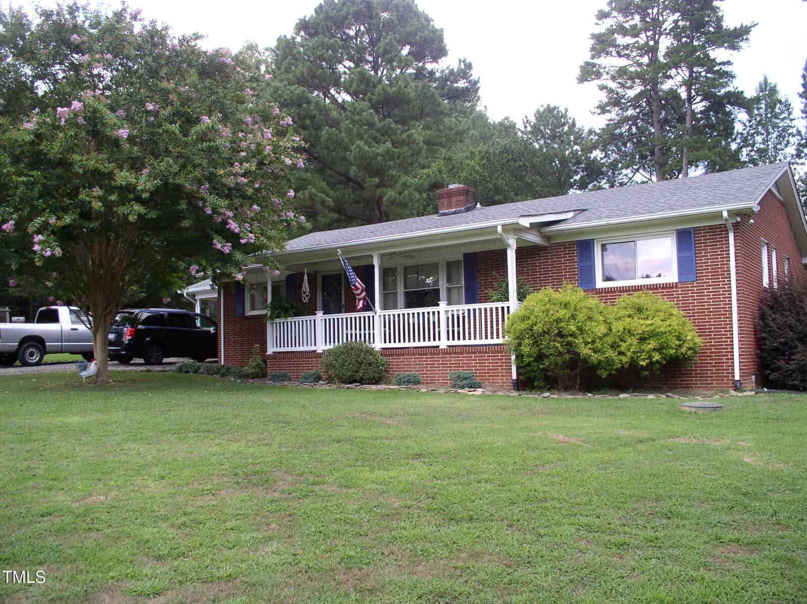 A single-story brick house at 6708 Old Roxboro Road, Oxford, features a covered front porch, blue shutters, and an American flag. The grassy yard has a large tree on the left, several bushes near the house, and two vehicles parked to the left.