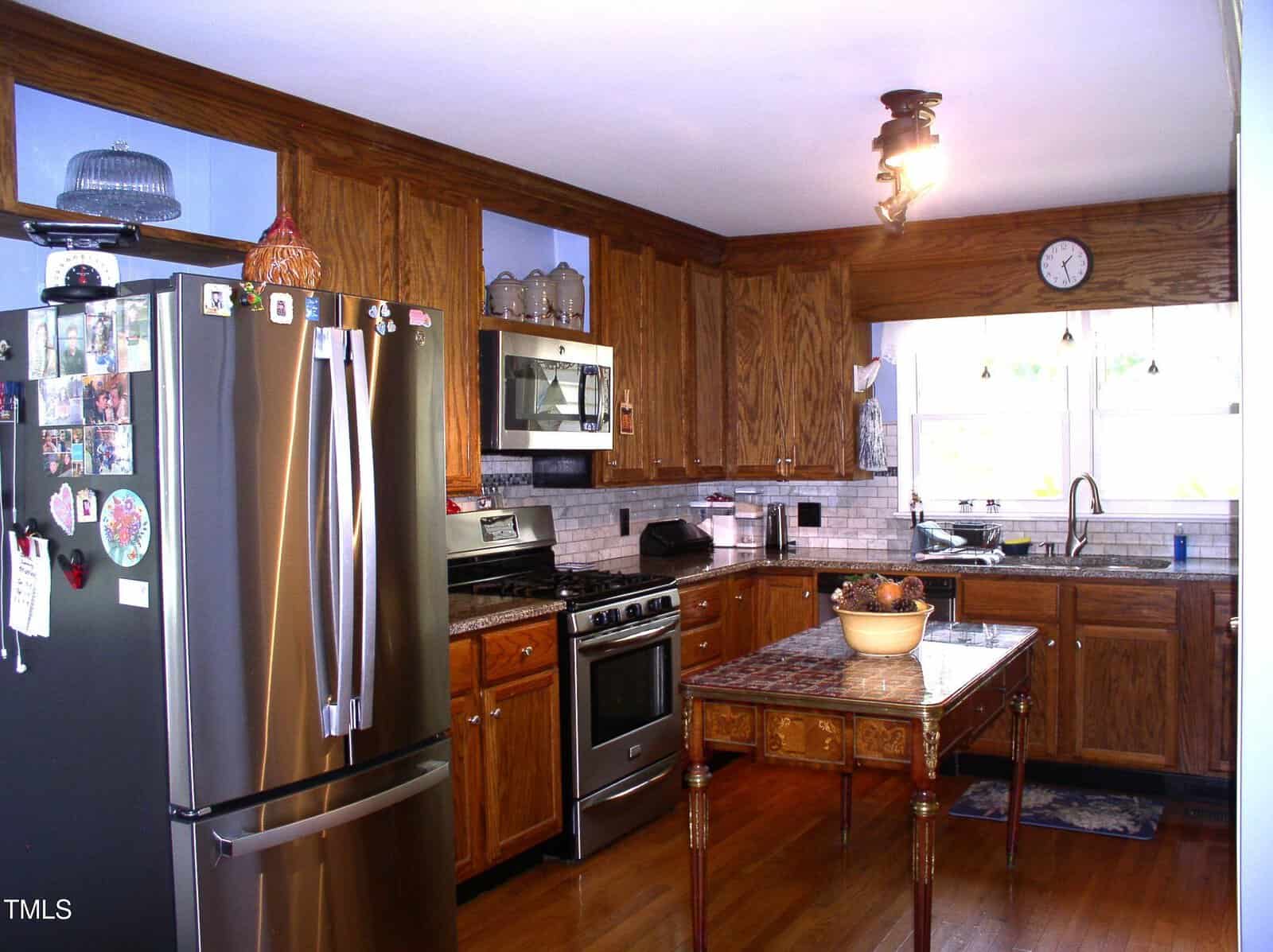 Kitchen with wood cabinets, stainless steel appliances, a refrigerator covered in magnets, a gas stove, a microwave, a double sink under two windows, and a wooden table with a bowl of fruit in the center.