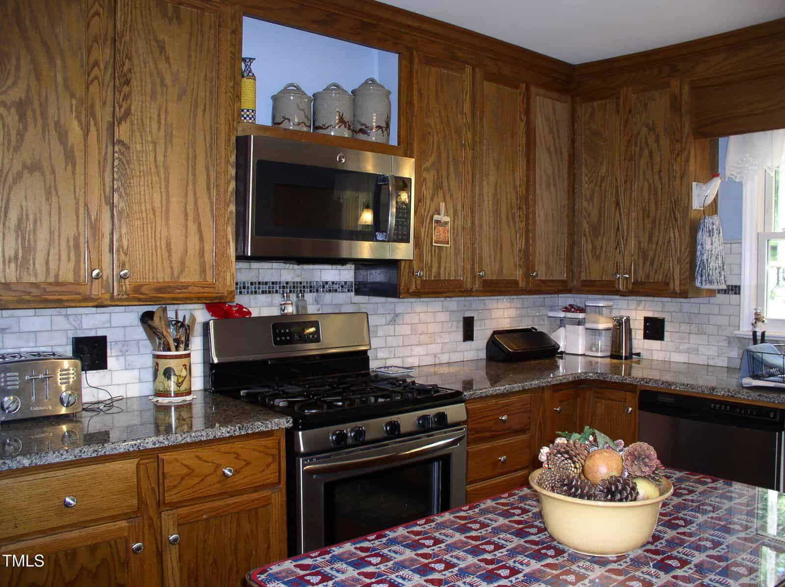 A kitchen with wooden cabinets, a stainless steel gas stove, microwave, and dishwasher, gray granite countertops, a tile backsplash, and small appliances along the counter. A fruit bowl sits on a table with a patterned tablecloth.