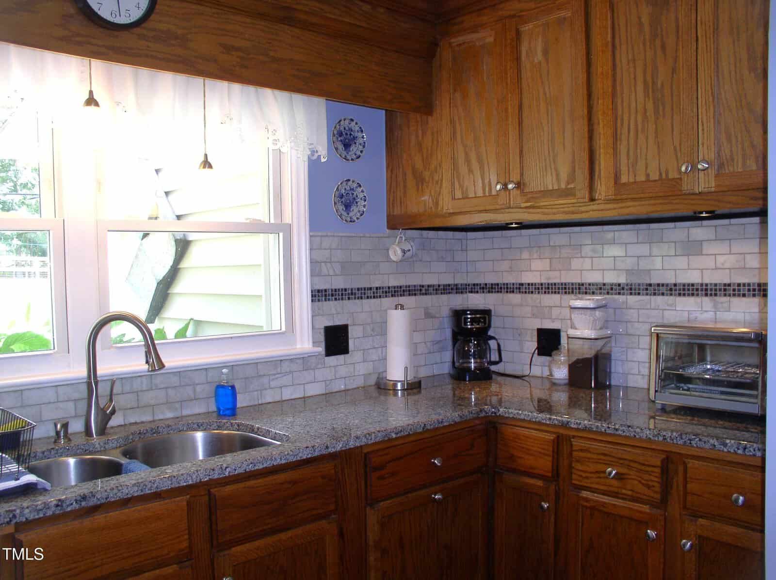 A kitchen corner with wooden cabinets, a granite countertop, a double sink, a window with a white curtain, a coffee maker, a toaster oven, a canister, and a blue soap dispenser. White subway tile backsplash.