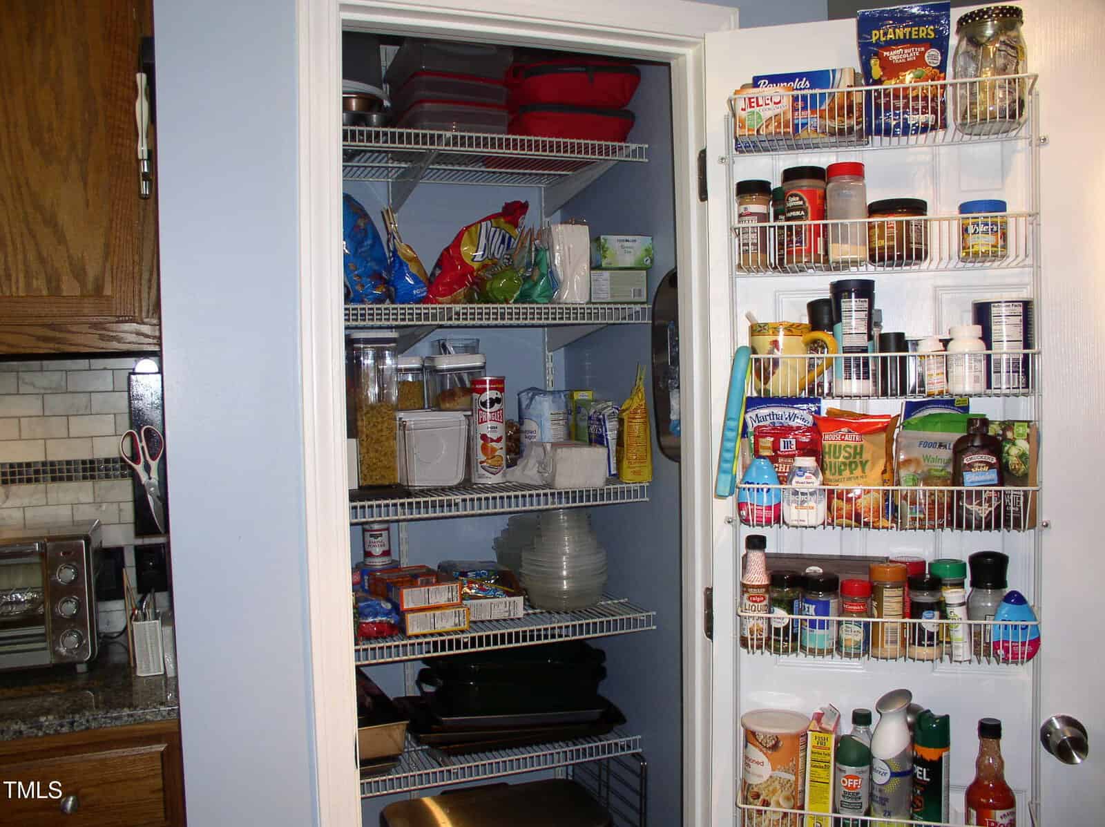 A pantry with wire shelves holds jars, boxes, canned goods, dishes, and baking pans. The door racks display spices, condiments, and snacks. A toaster oven is visible on the adjacent kitchen counter.