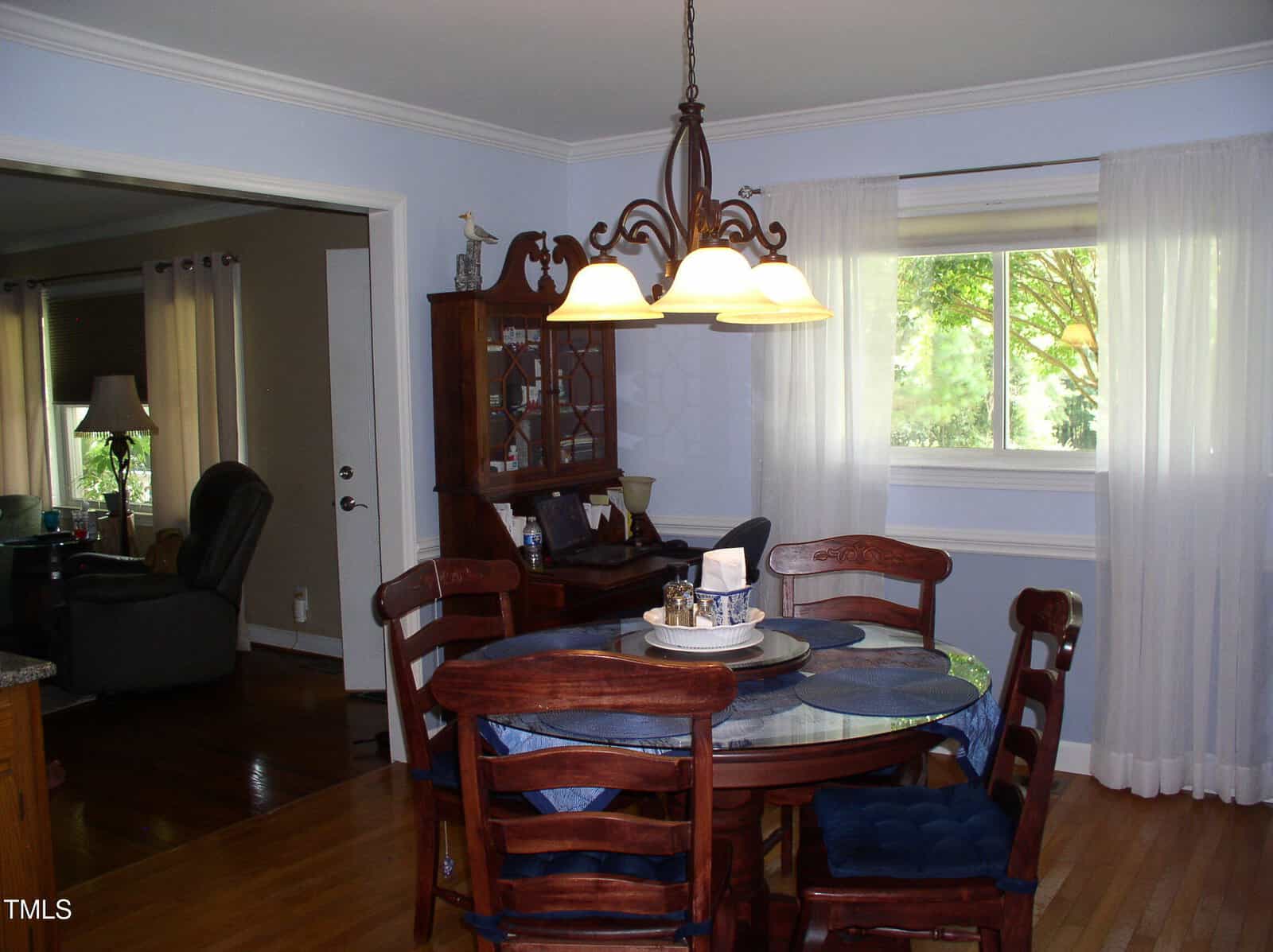 A dining area with a round wooden table and four chairs, set with placemats and a centerpiece. A chandelier hangs above. There is a hutch, a window with sheer curtains, and an adjacent living room visible.