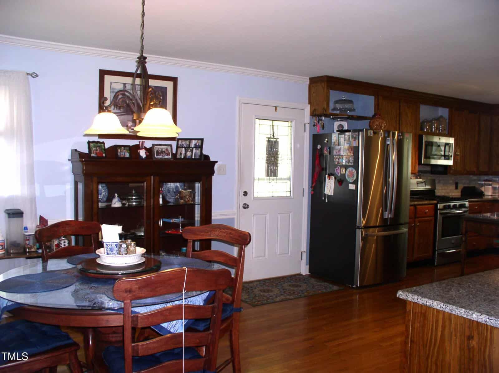 A kitchen and dining area with wooden cabinets, stainless steel appliances, a round dining table with four chairs, a glass-front cabinet, and a white door with a decorative window.
