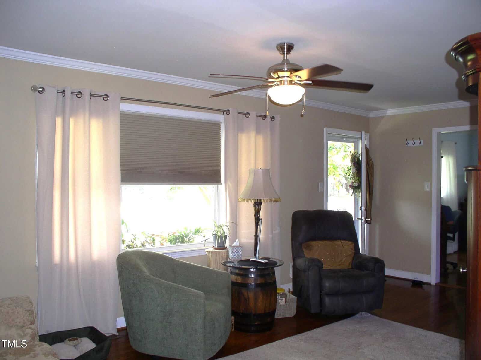 A living room with beige walls, a ceiling fan with a light, two armchairs, a barrel used as a side table, a floor lamp, and large windows with curtains. Hardwood floor and a door to the outside are visible.